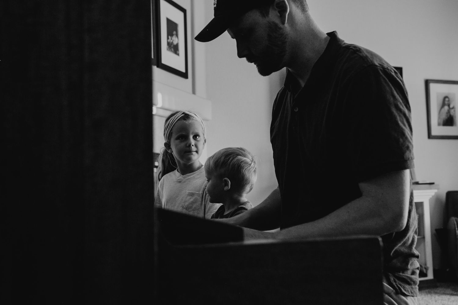 A black and white candid moment captured indoors with figures gathered around a piano.