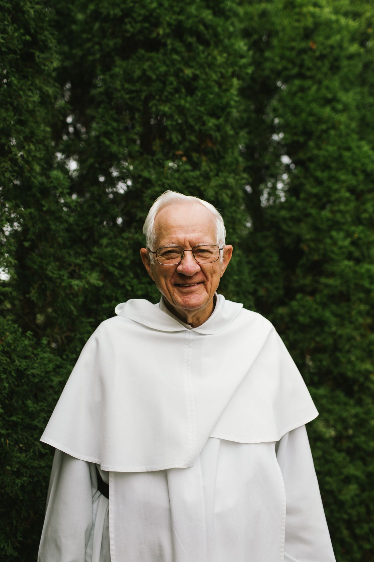An elderly man in white robes smiles warmly against a green leafy background.