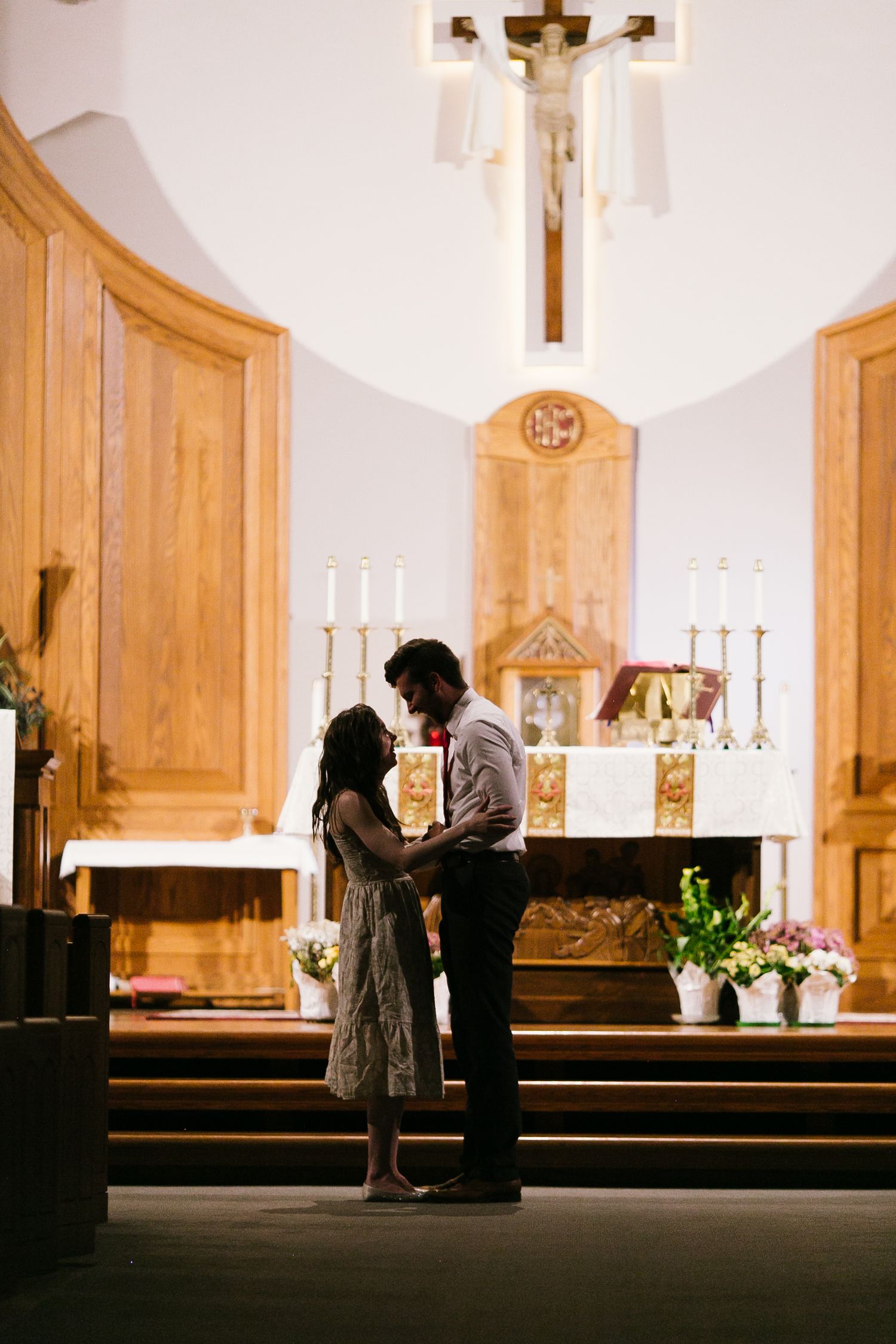 A couple shares an intimate moment in a church sanctuary with wooden pews and a crucifix in the background.