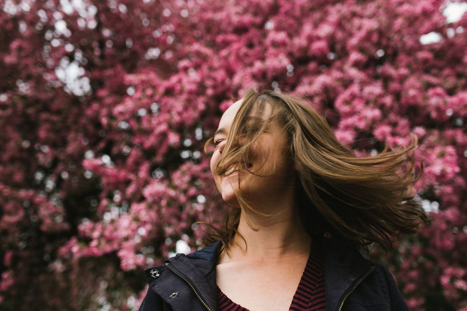 A person with windswept hair stands against blooming pink cherry blossoms.