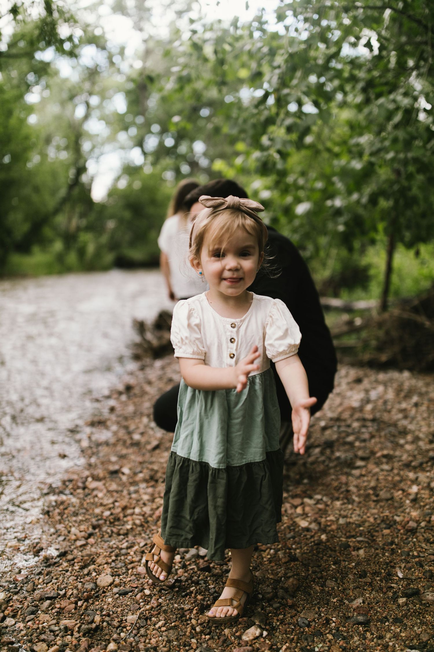 A child in a tiered dress stands on a dirt path surrounded by lush green foliage.