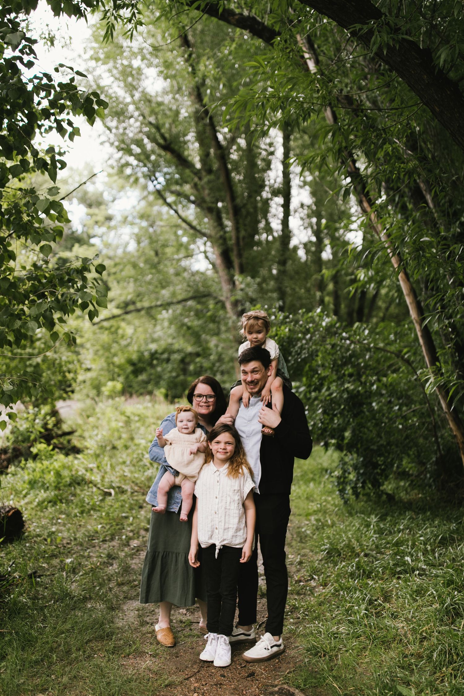 A family poses together on a wooded trail surrounded by green trees and natural scenery.