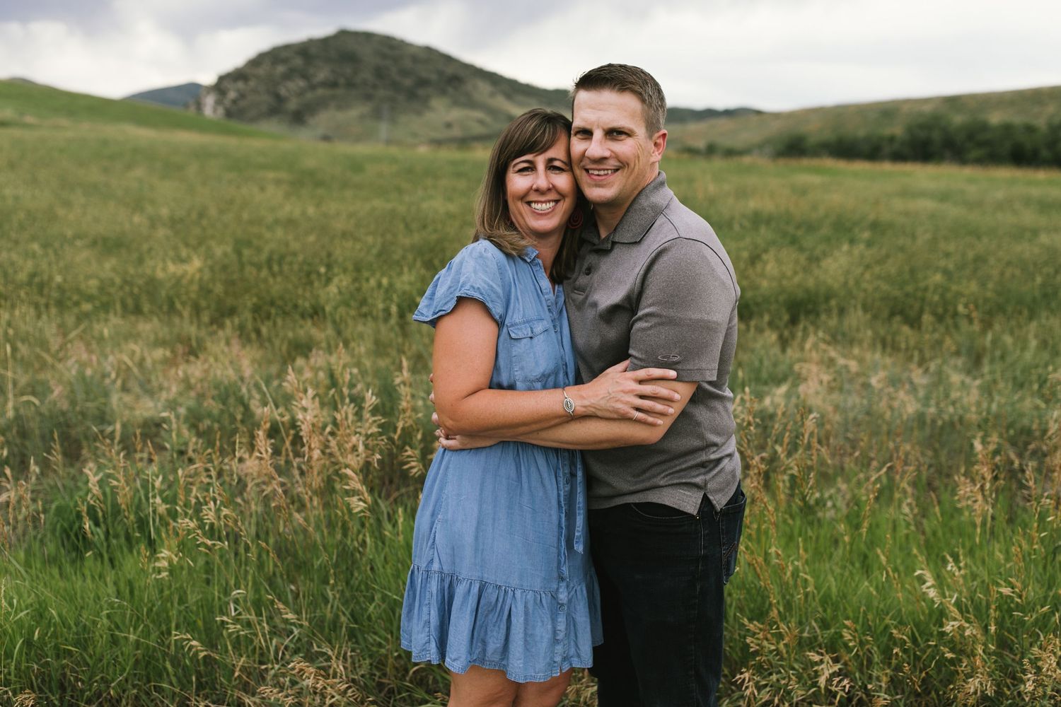 A couple embraces in a grassy meadow with rolling hills in the background.