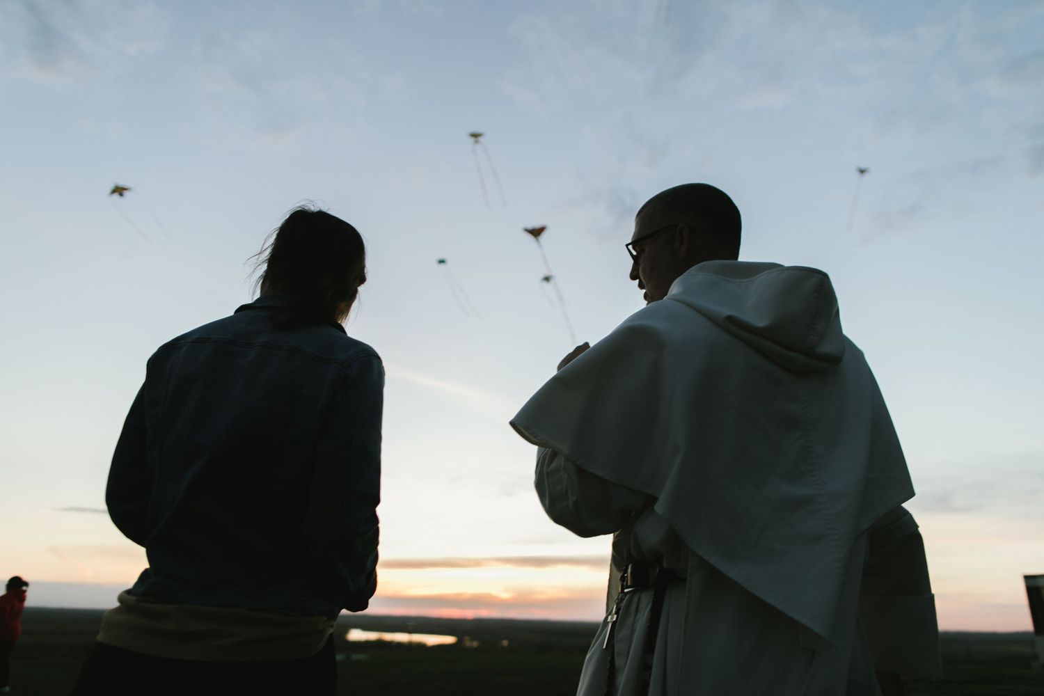 Two silhouetted figures watch birds flying in the sky during a peaceful sunset.