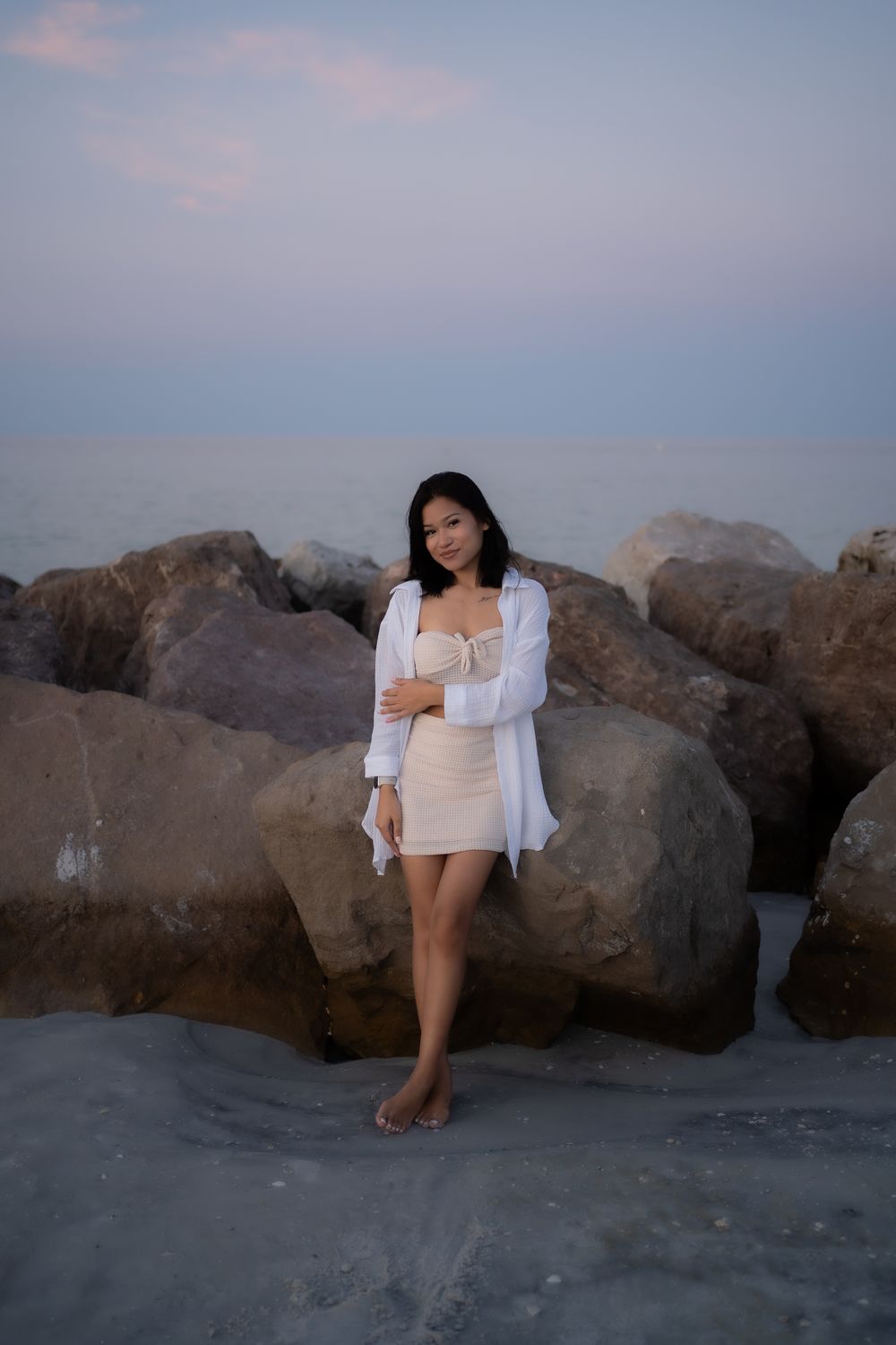 A person in a white button-down shirt and light dress stands barefoot against large coastal rocks at dusk.