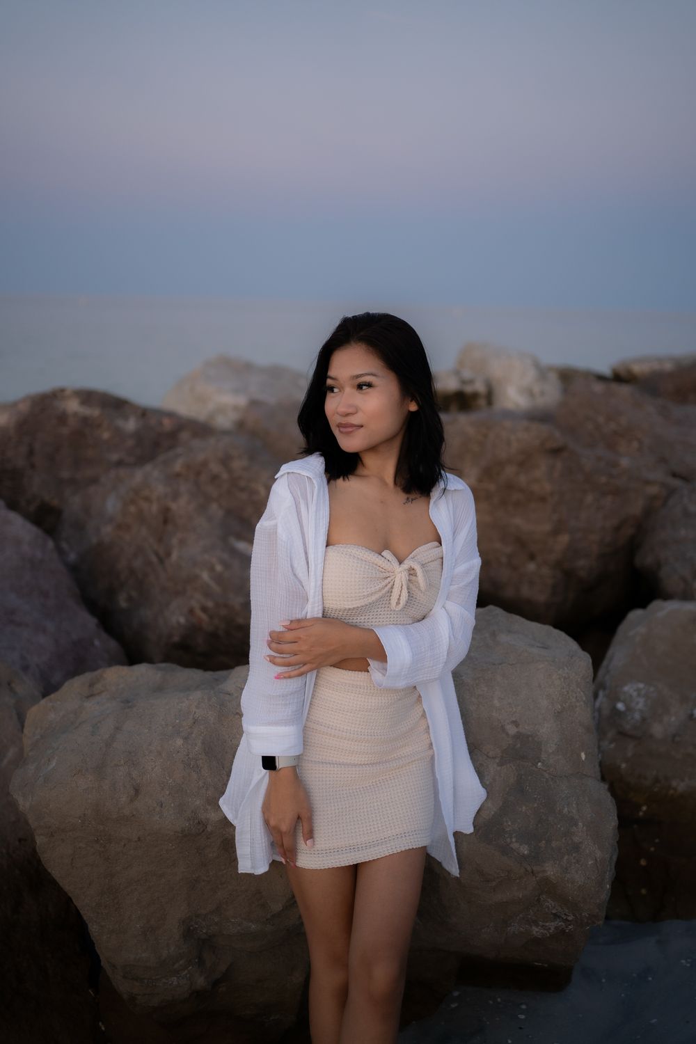 A person in a beige dress and white cardigan poses against large rocks during sunset at the beach.