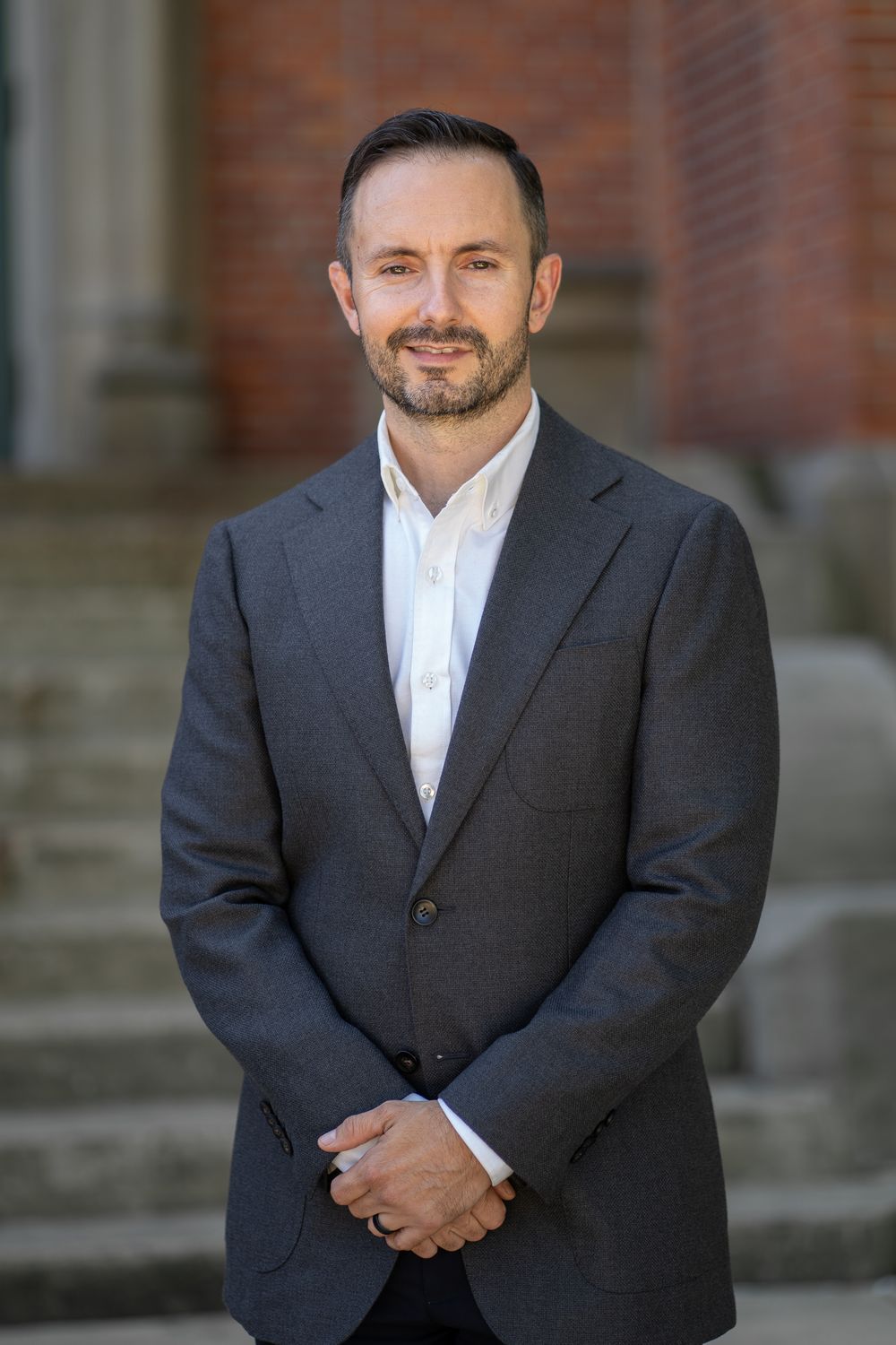 Professional business portrait of person in dark suit and white shirt standing outdoors by brick building with stone steps.