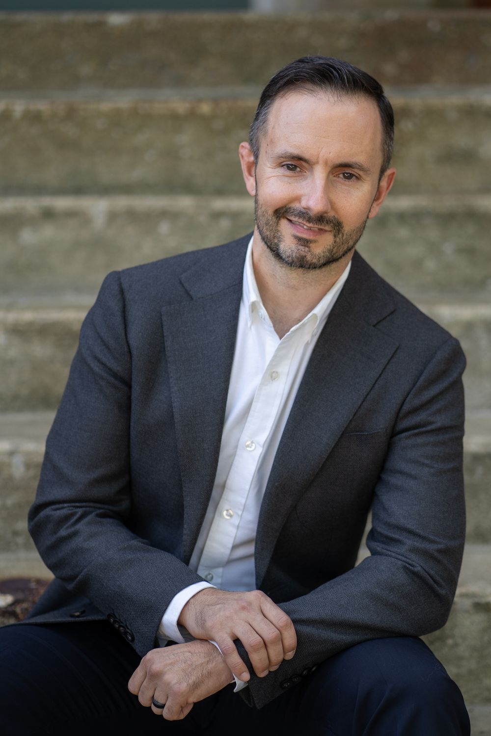 Professional headshot of a well-groomed man in dark suit and white shirt seated on stone steps outdoors.