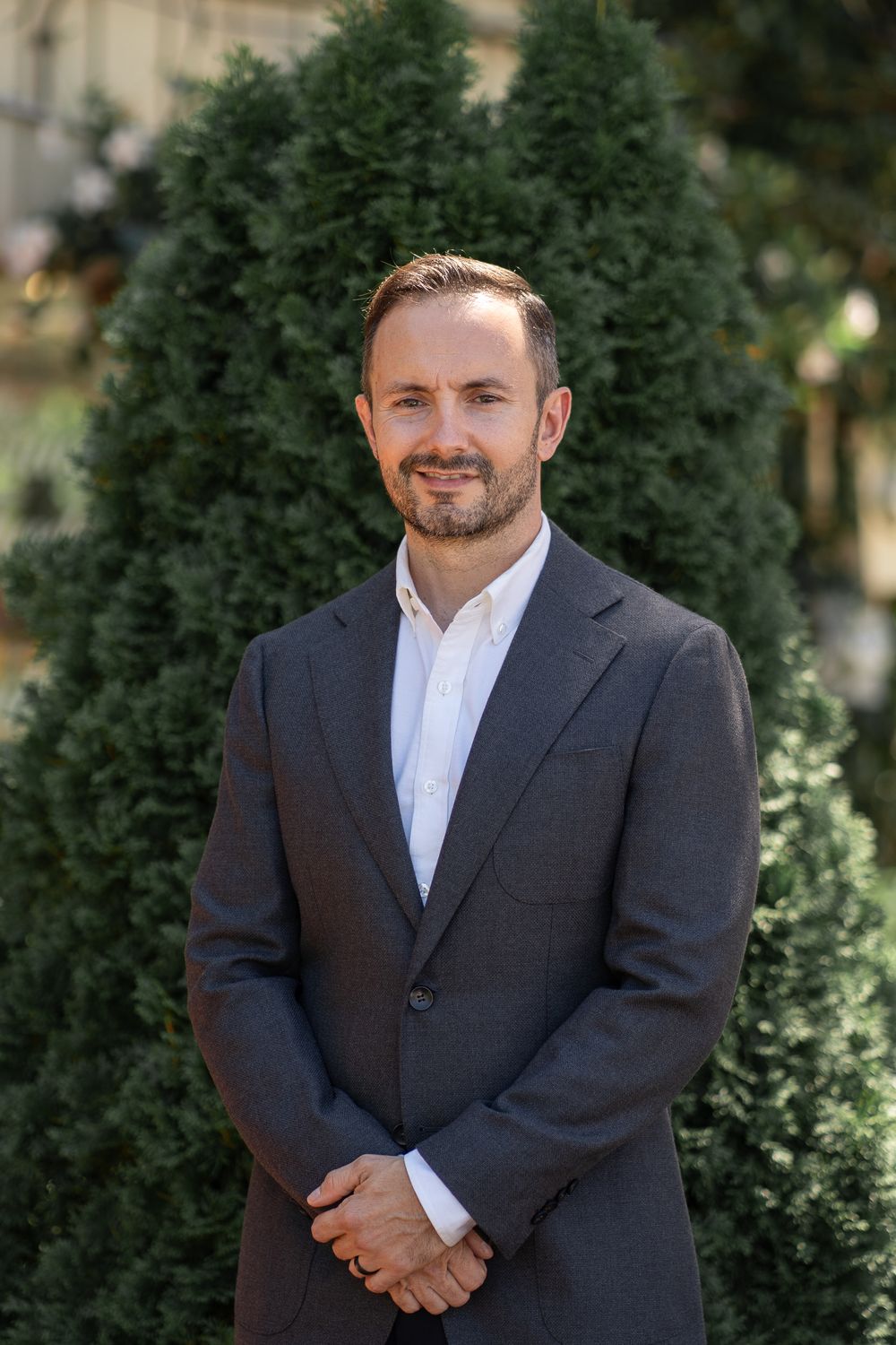 Professional portrait of a businessman wearing a dark suit and white shirt standing in front of evergreen trees outdoors.