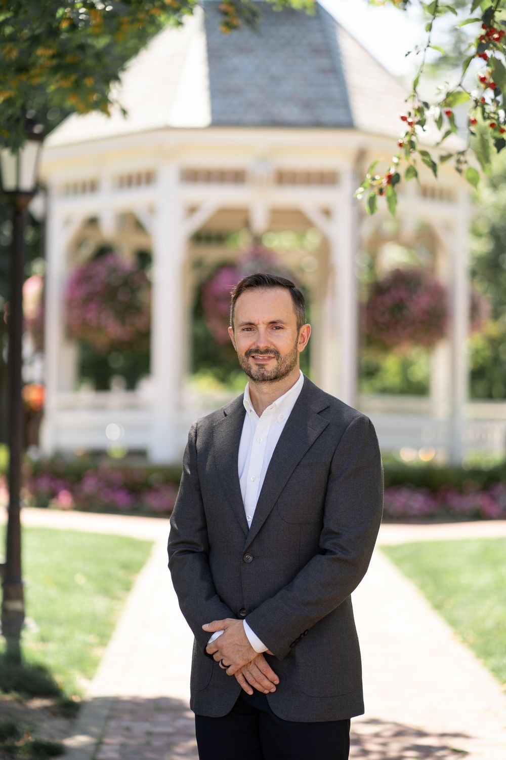 A professional in a dark suit jacket and white shirt stands in front of a white gazebo surrounded by colorful flowers.
