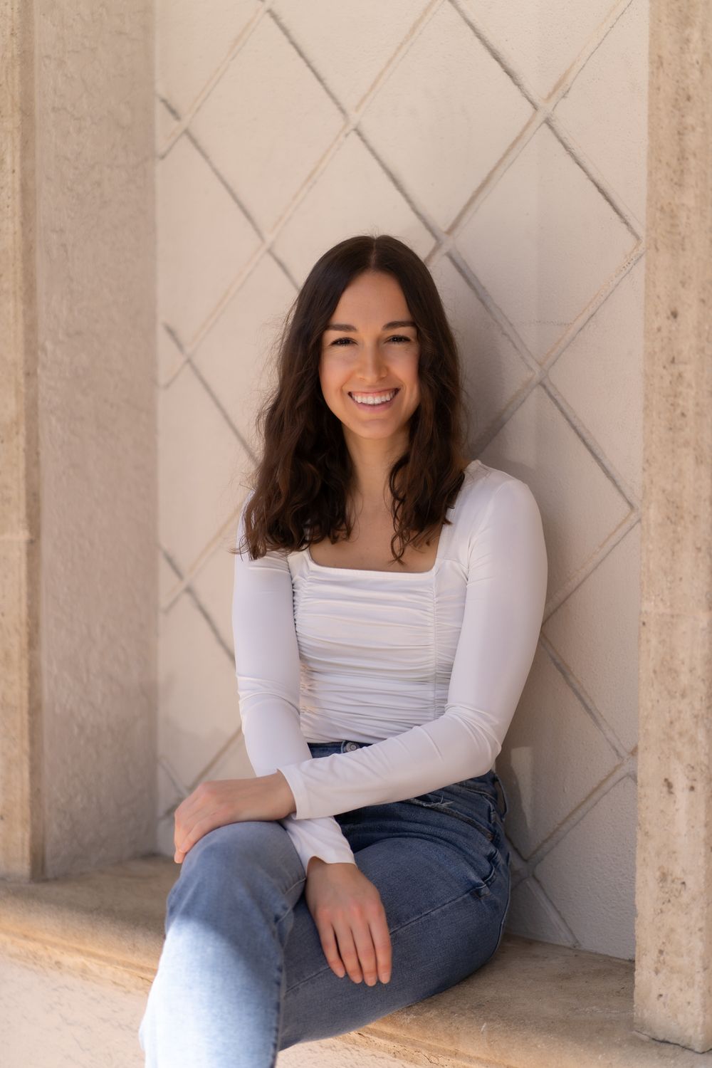 A smiling person wearing a white long-sleeve top and jeans sits casually against a geometric patterned wall.