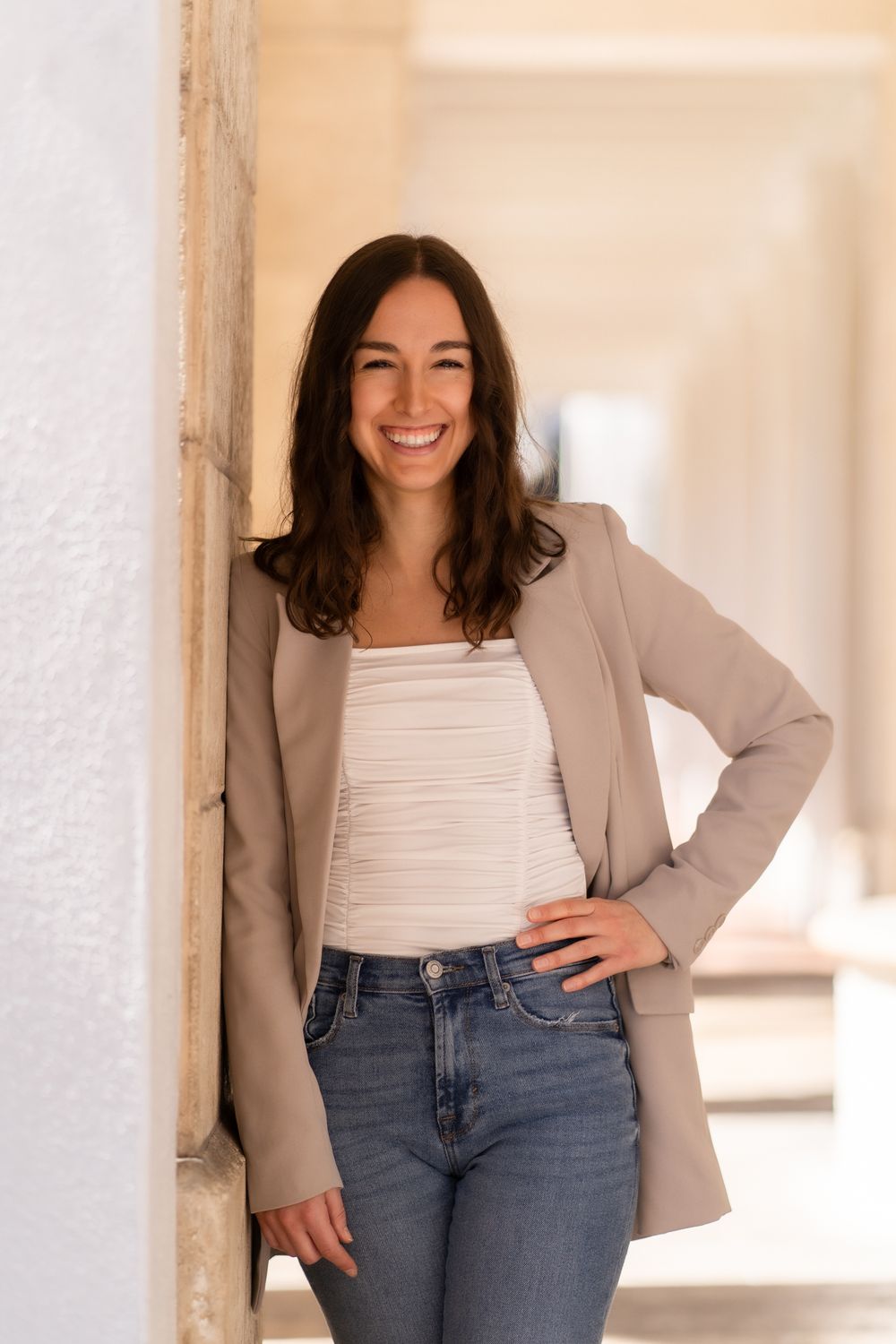 Professional portrait of a person in a beige blazer and white top leaning against a white wall in a bright hallway.