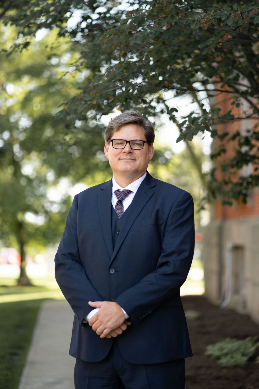 Professional portrait series of person in navy suit and glasses against blurred outdoor background with trees.