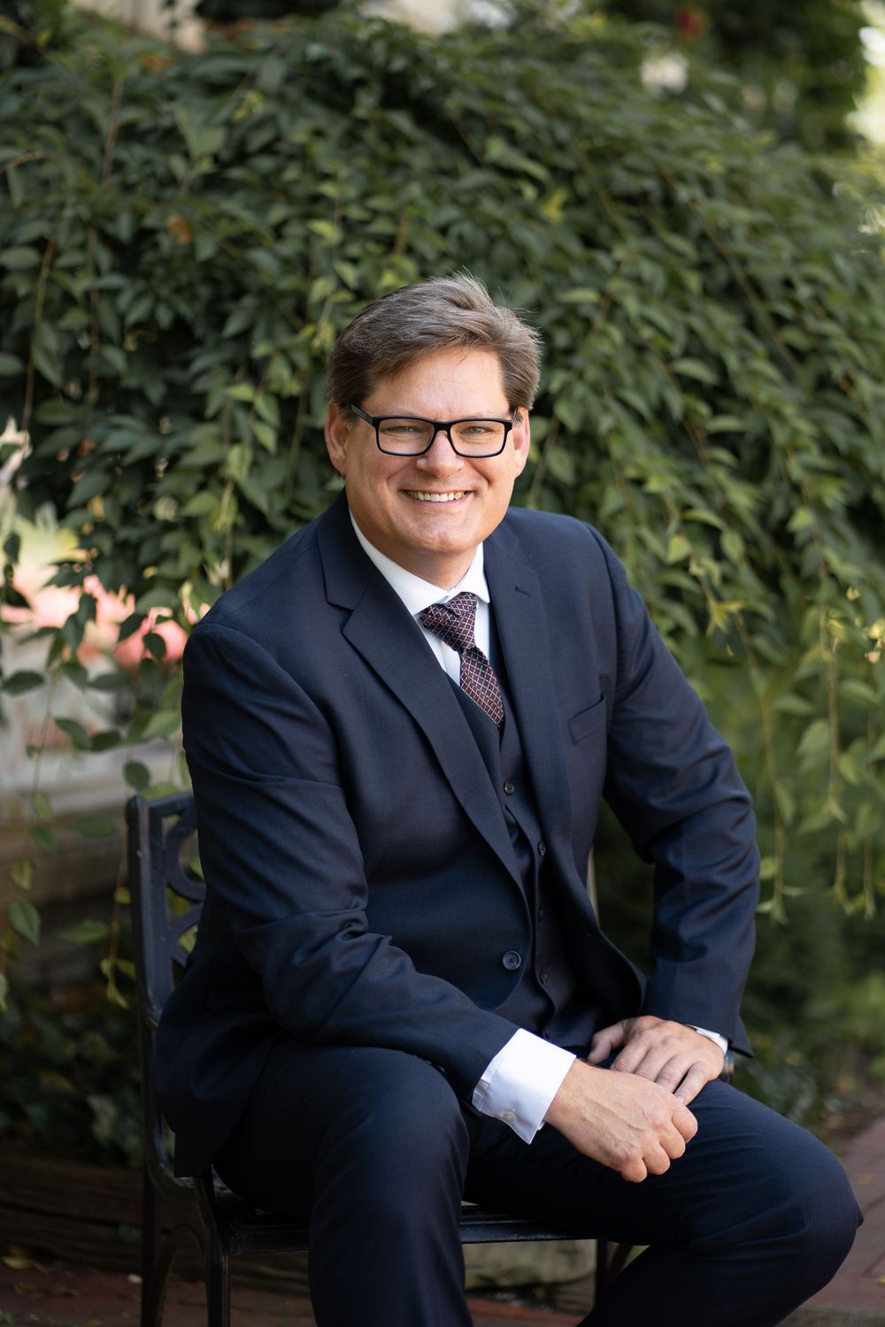Professional portrait of someone in a dark business suit and glasses sitting on a bench with greenery in background.