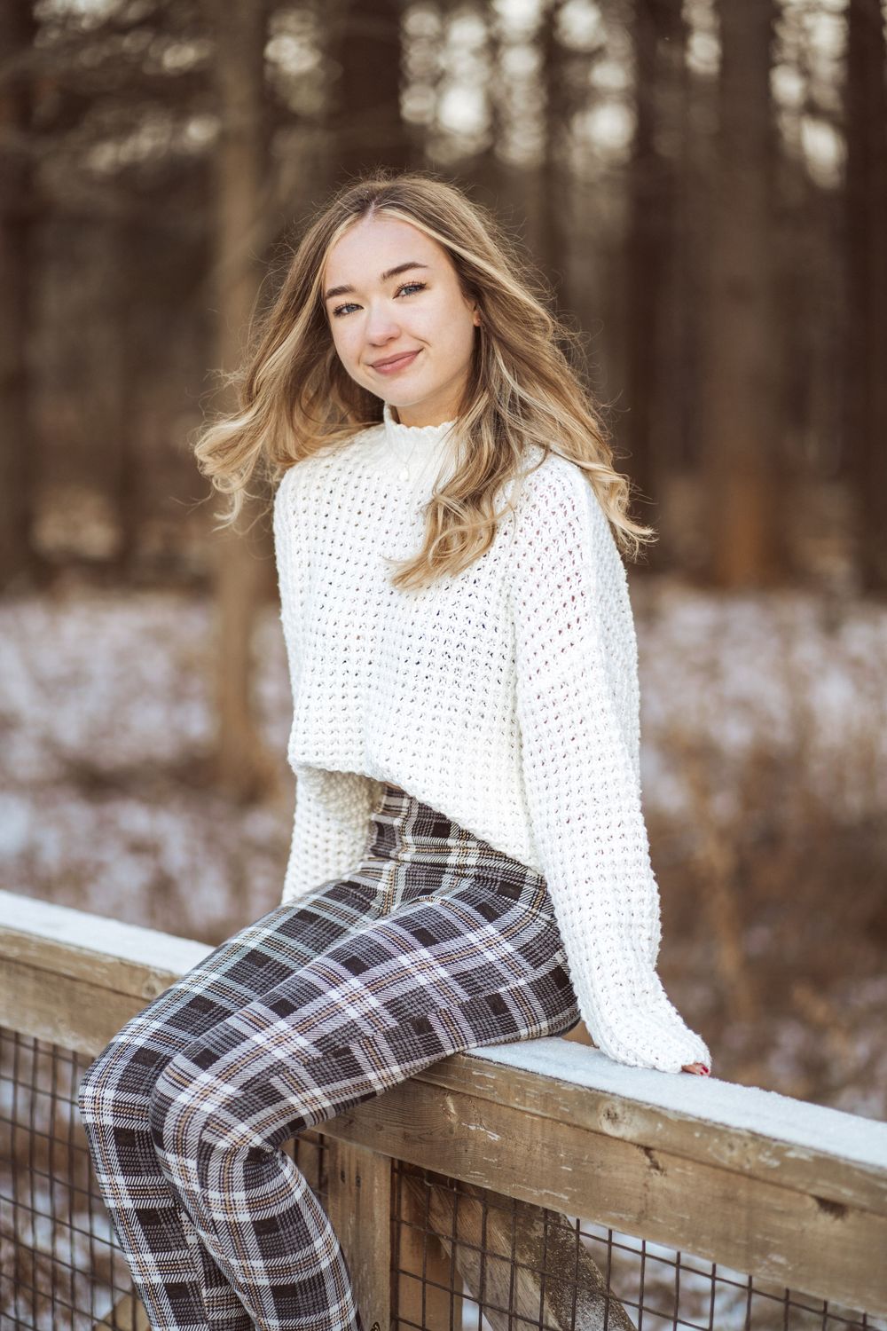 Stylish winter outfit featuring white sweater and plaid pants photographed in snowy outdoor setting on wooden railing.