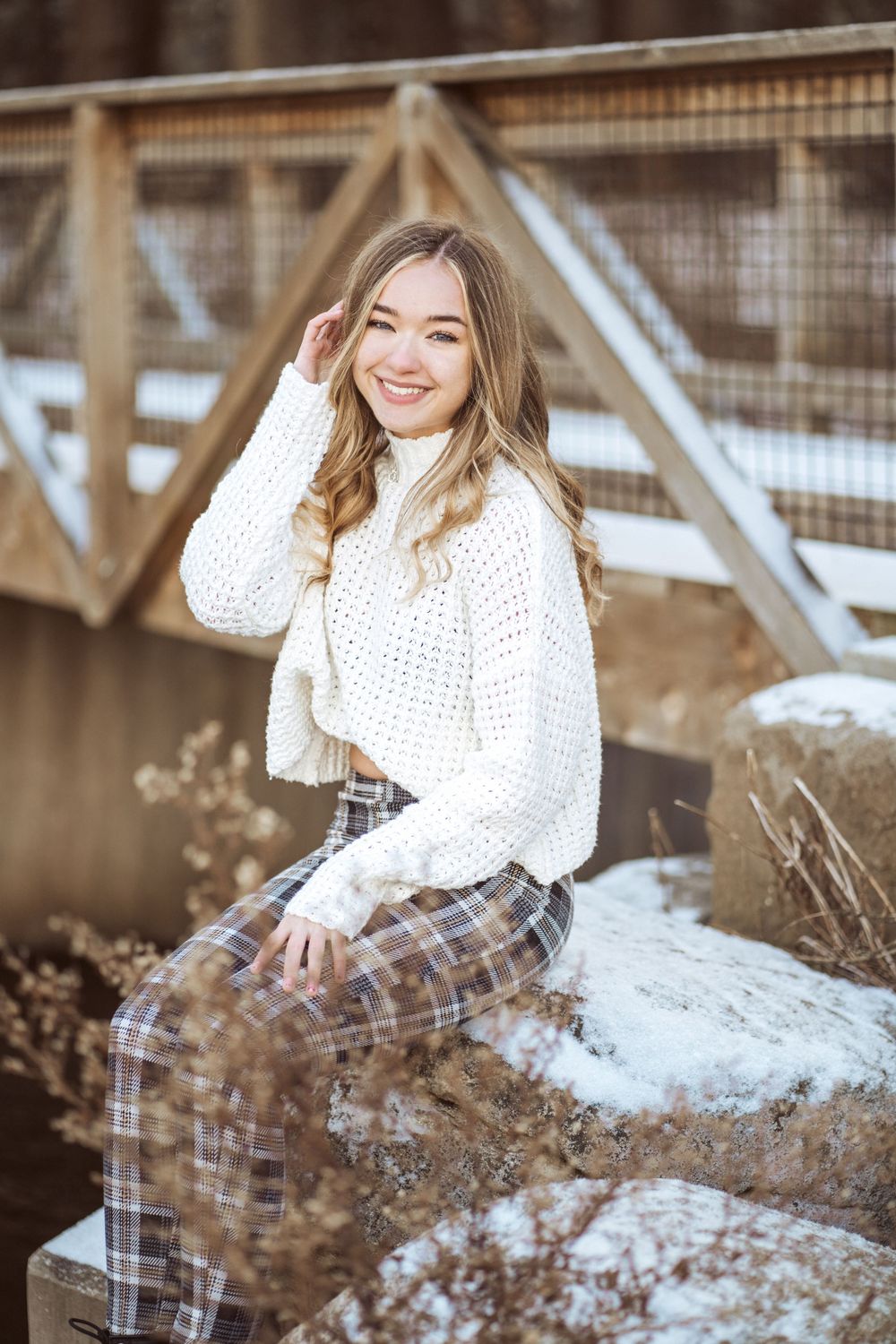 Someone in a cozy white sweater and plaid pants sits on a snowy fence near a wooden bridge structure.