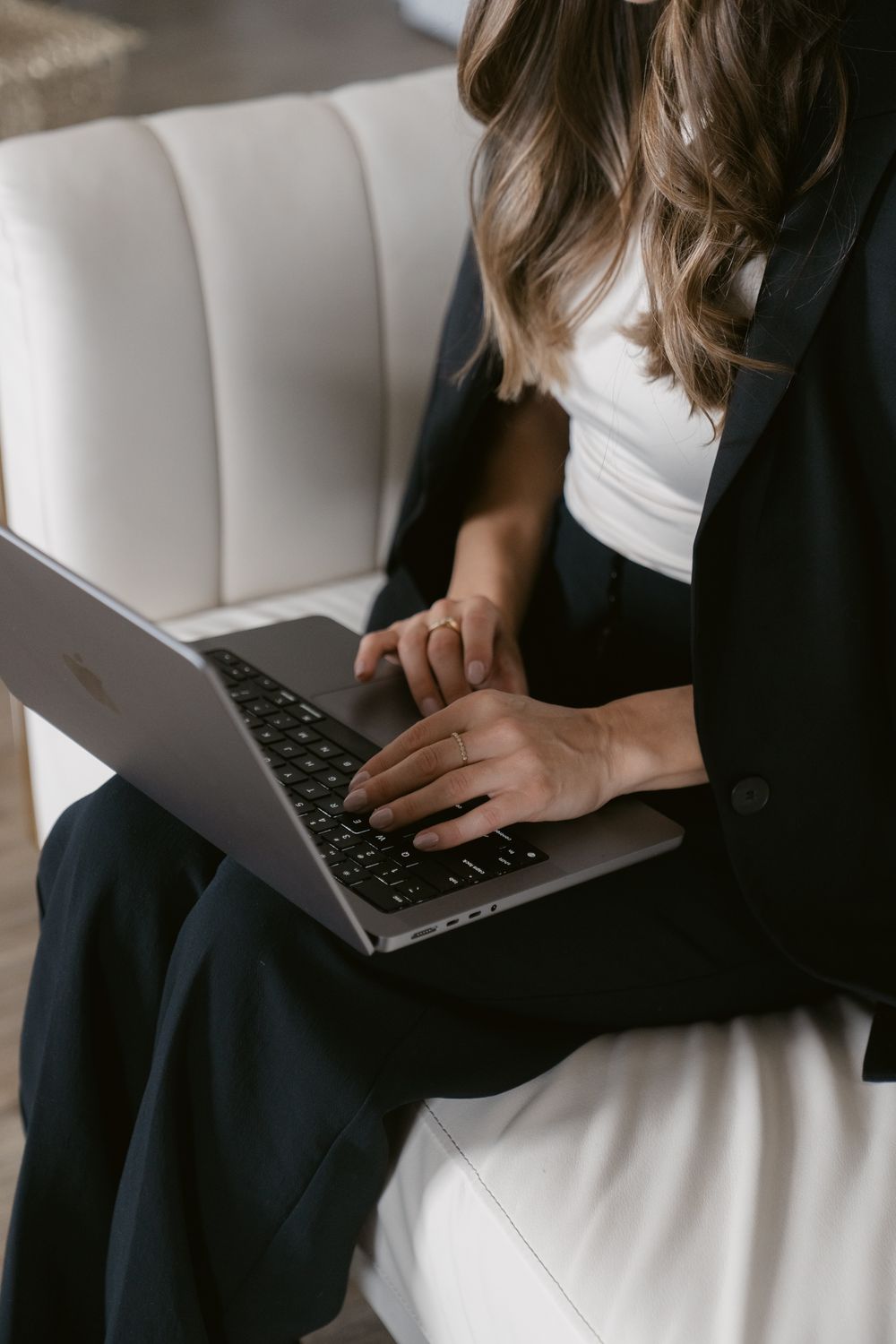 Person in business attire working on laptop while sitting on cream-colored upholstered seating.