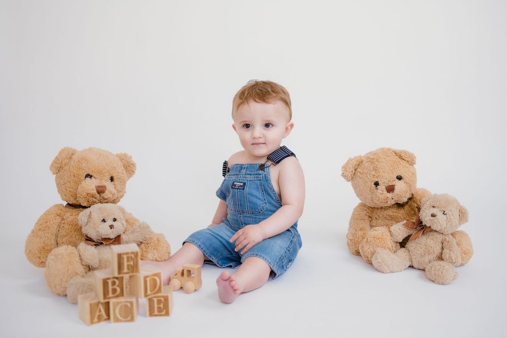 Adorable toddler in denim overalls posed between two teddy bears with wooden letter blocks.