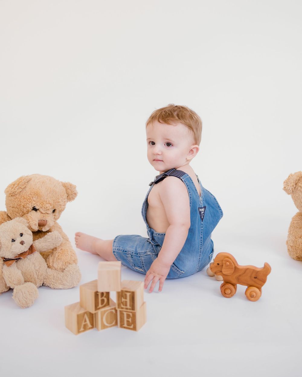 Cute baby in denim overalls sitting with teddy bears and wooden alphabet blocks on white background.