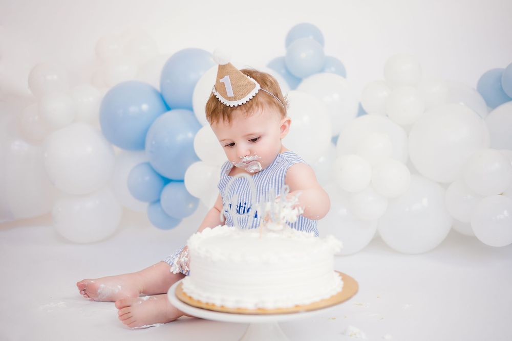 Baby in blue outfit reaching toward first birthday smash cake surrounded by blue and white balloons.