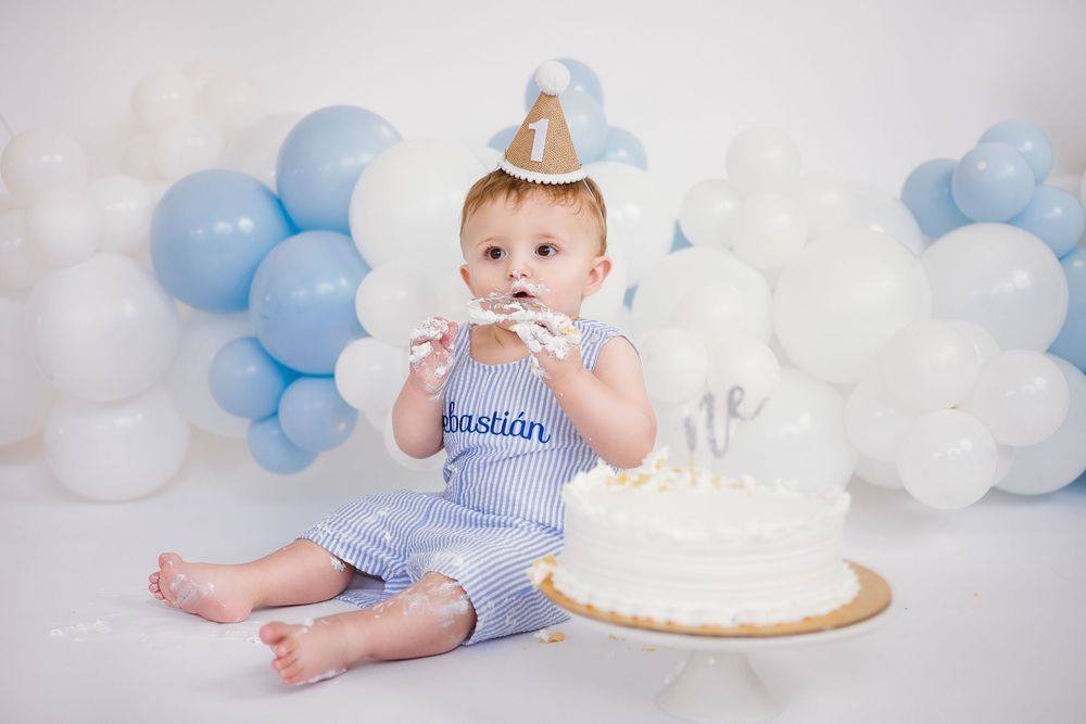 Baby boy named Sebastian eating first birthday smash cake with blue and white balloon backdrop.