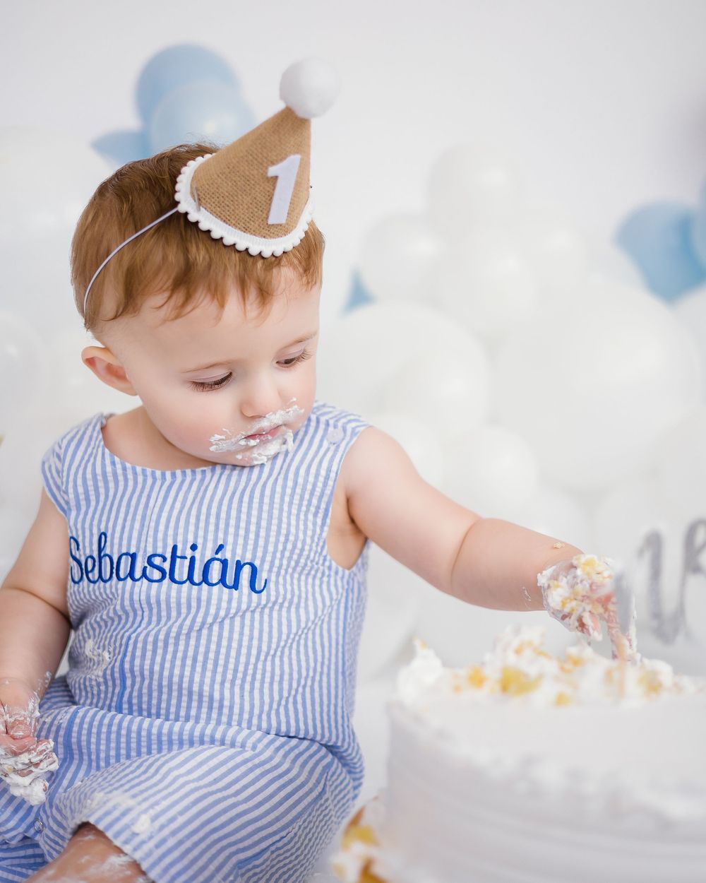 Baby Sebastian in striped outfit touching smash cake during first birthday celebration with balloons.