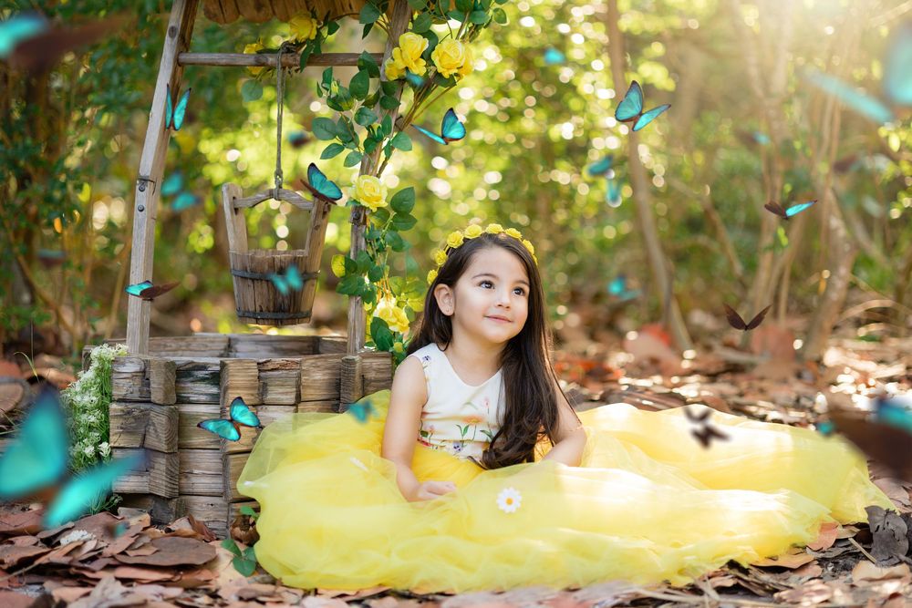 A little girl in a yellow tulle dress sits surrounded by colorful butterflies outdoors.