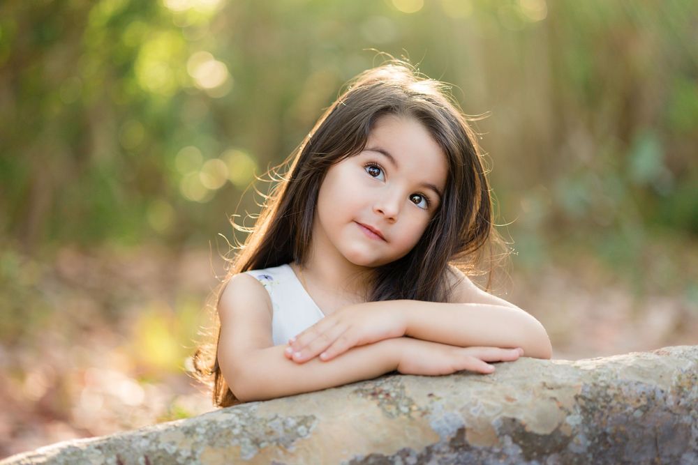 A pretty young girl with long dark hair rests her arms on a rock in a forest.
