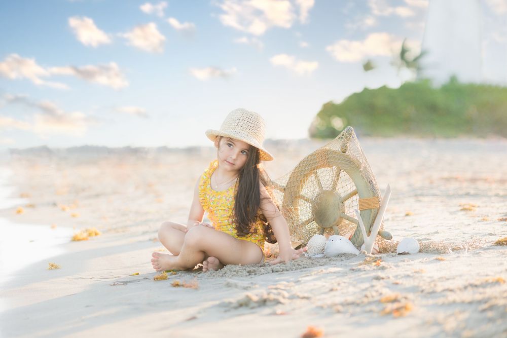 Young girl in yellow swimsuit and straw hat sitting on sandy beach next to decorative ship wheel.