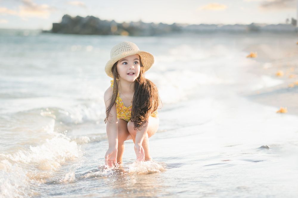 Adorable little girl in yellow floral swimsuit and straw hat plays in ocean waves on sunny beach.