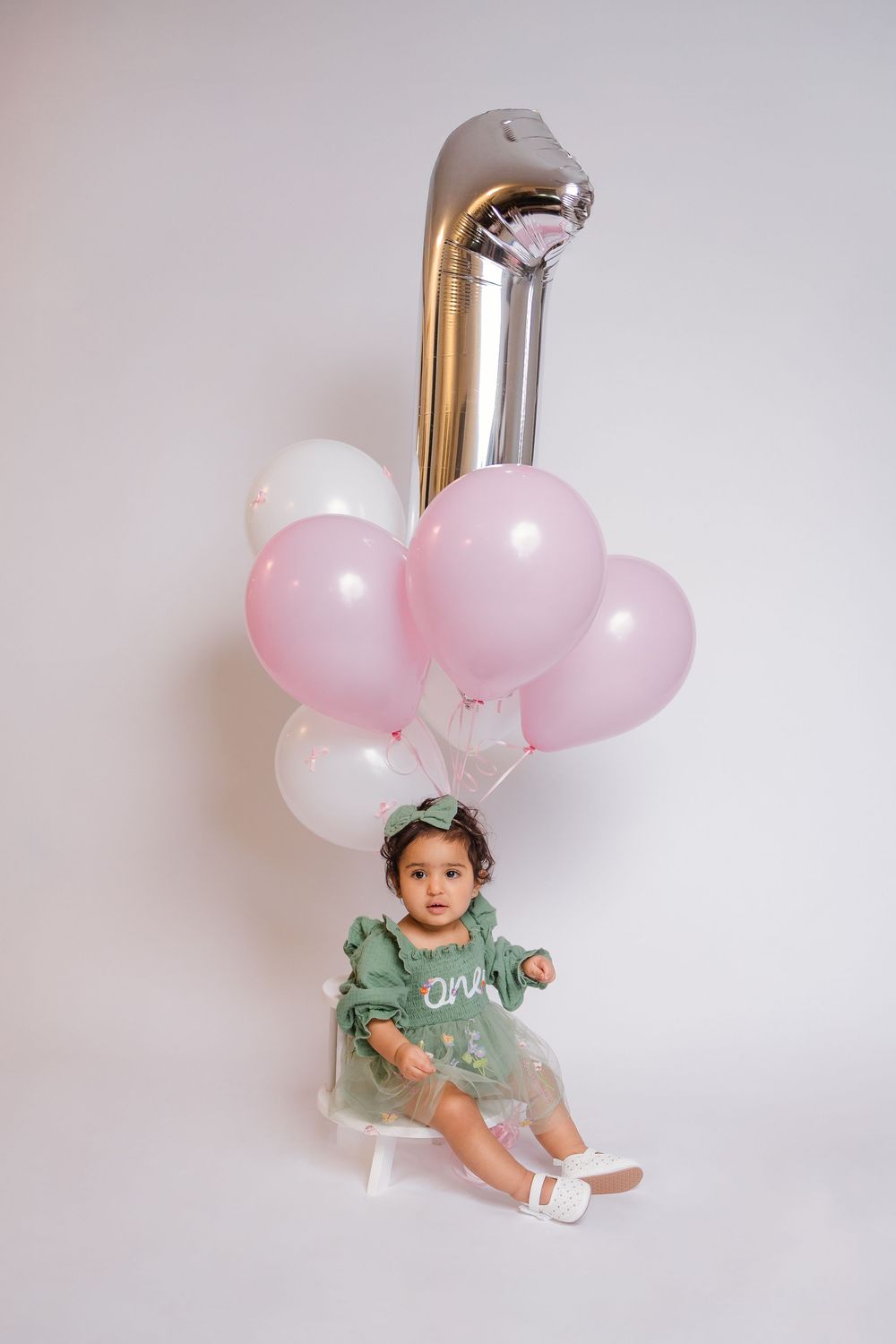 Baby girl in green one dress sits with pink and silver number one balloons for first birthday photos.