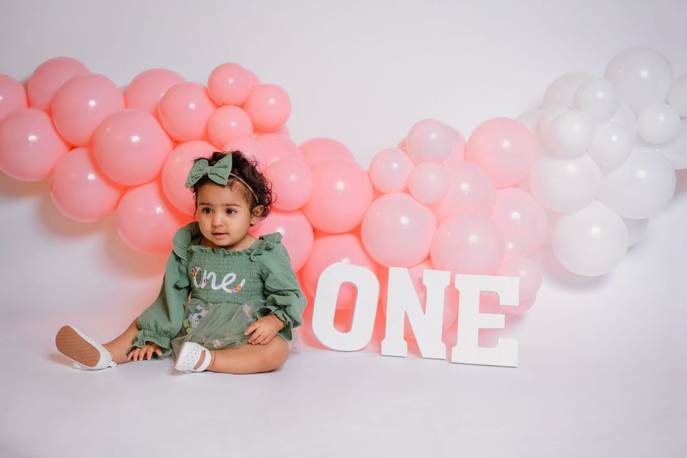 Smiling baby girl in green romper sits beside ONE sign surrounded by pink and white balloon garland.