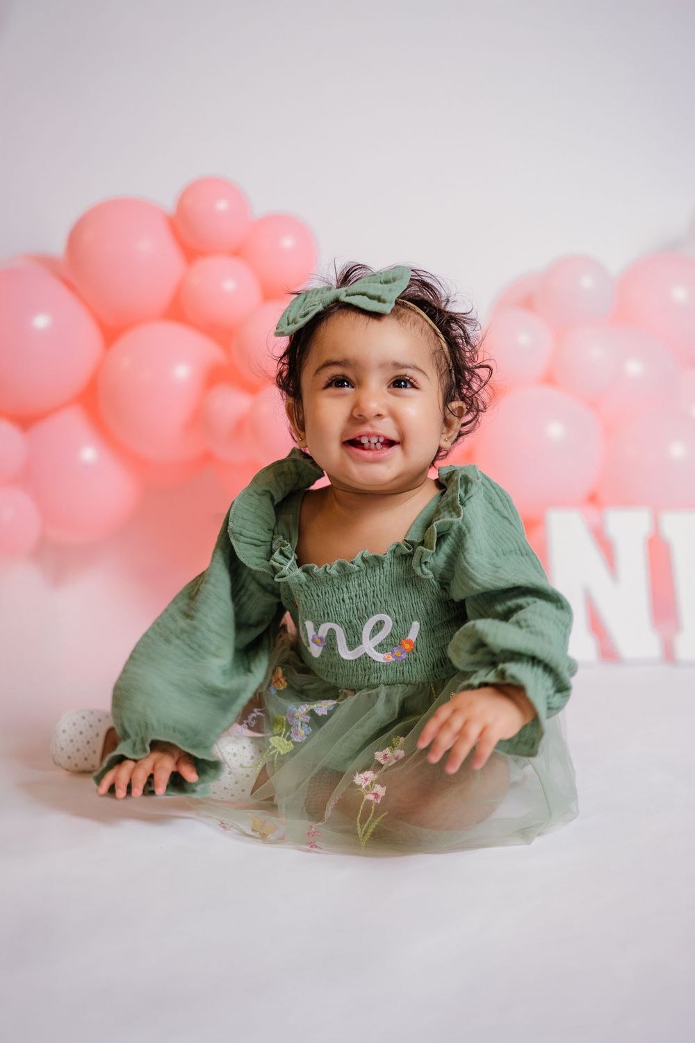 Happy one year old baby girl in green dress laughs in front of pink balloon backdrop for birthday photos.