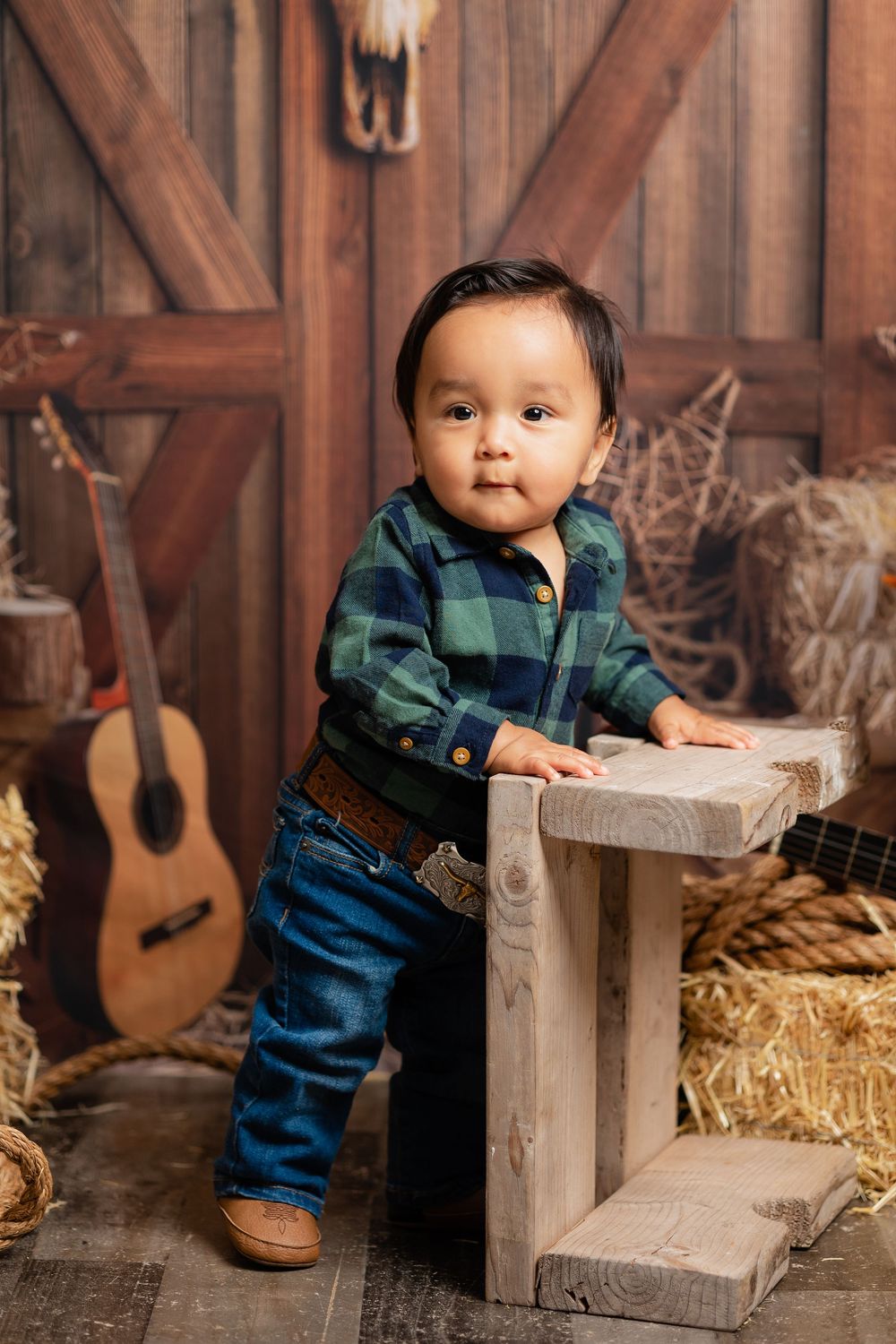 Adorable baby boy in plaid shirt and jeans poses in rustic western barn studio with hay bales and guitar.