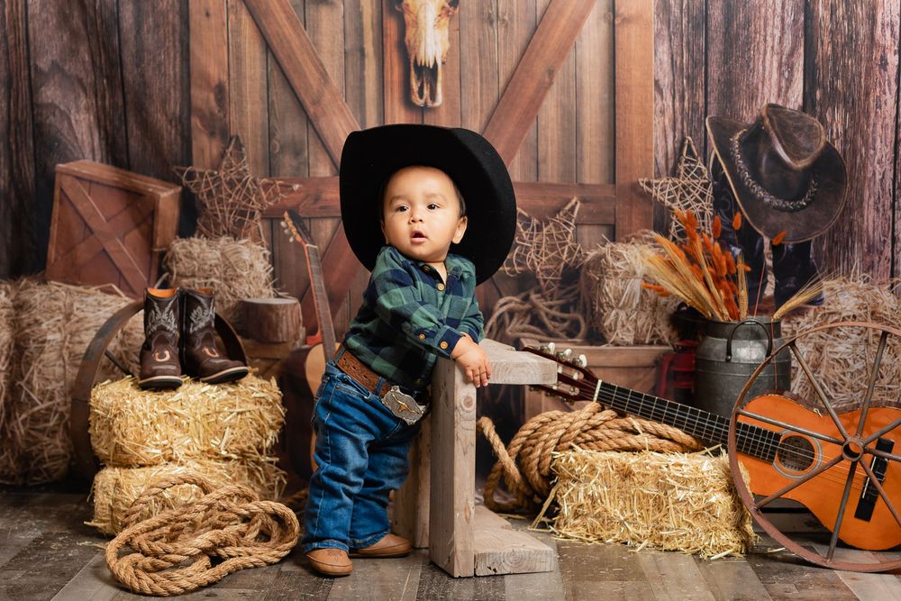 Cute toddler boy wearing oversized black cowboy hat poses in western barn studio with hay bales and guitar.