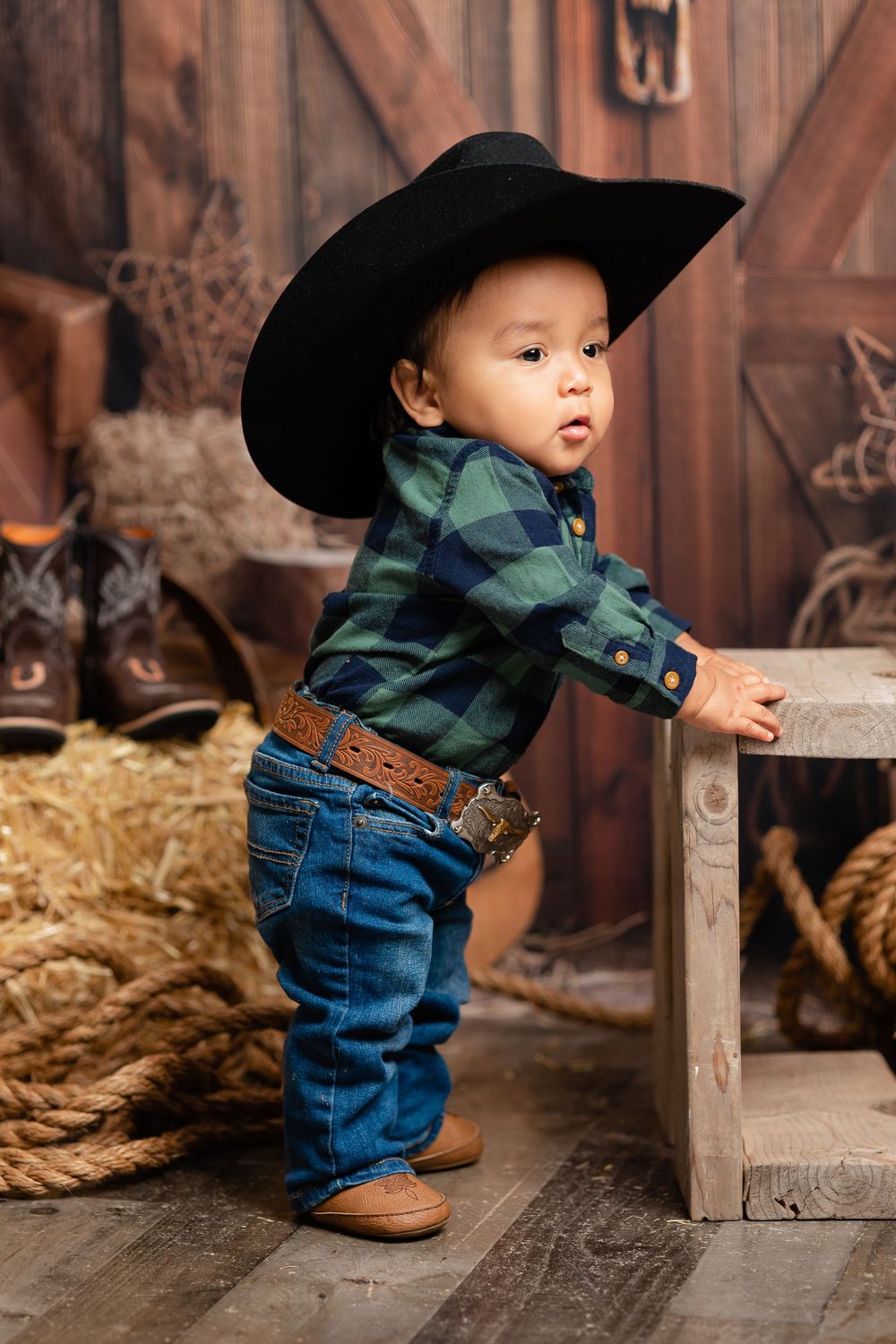 Sweet baby boy in black cowboy hat and plaid shirt stands by wooden crate in rustic western barn setting.