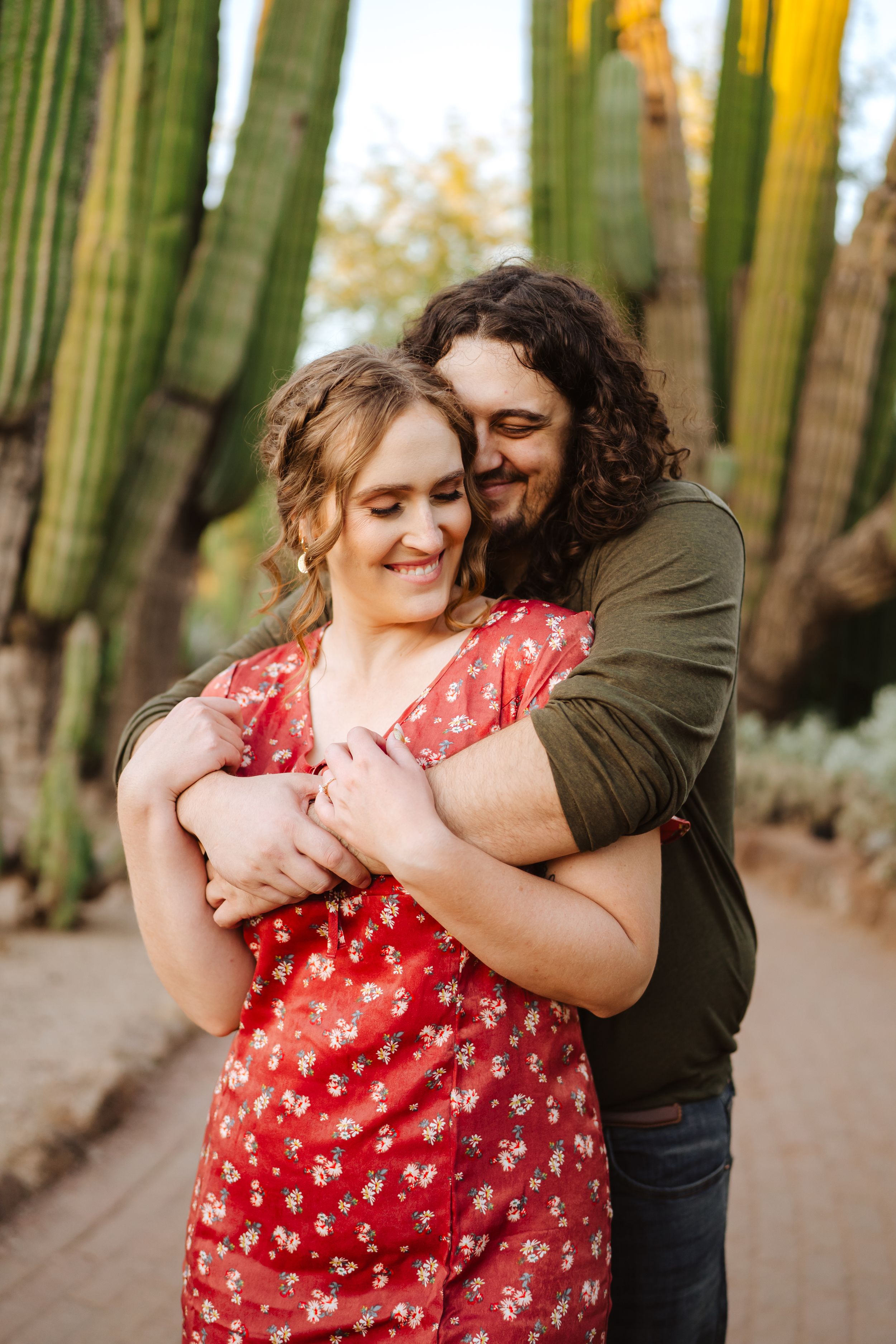 A couple embraces lovingly in a saguaro cactus-lined pathway while wearing red floral dress and olive green top during their engagement.