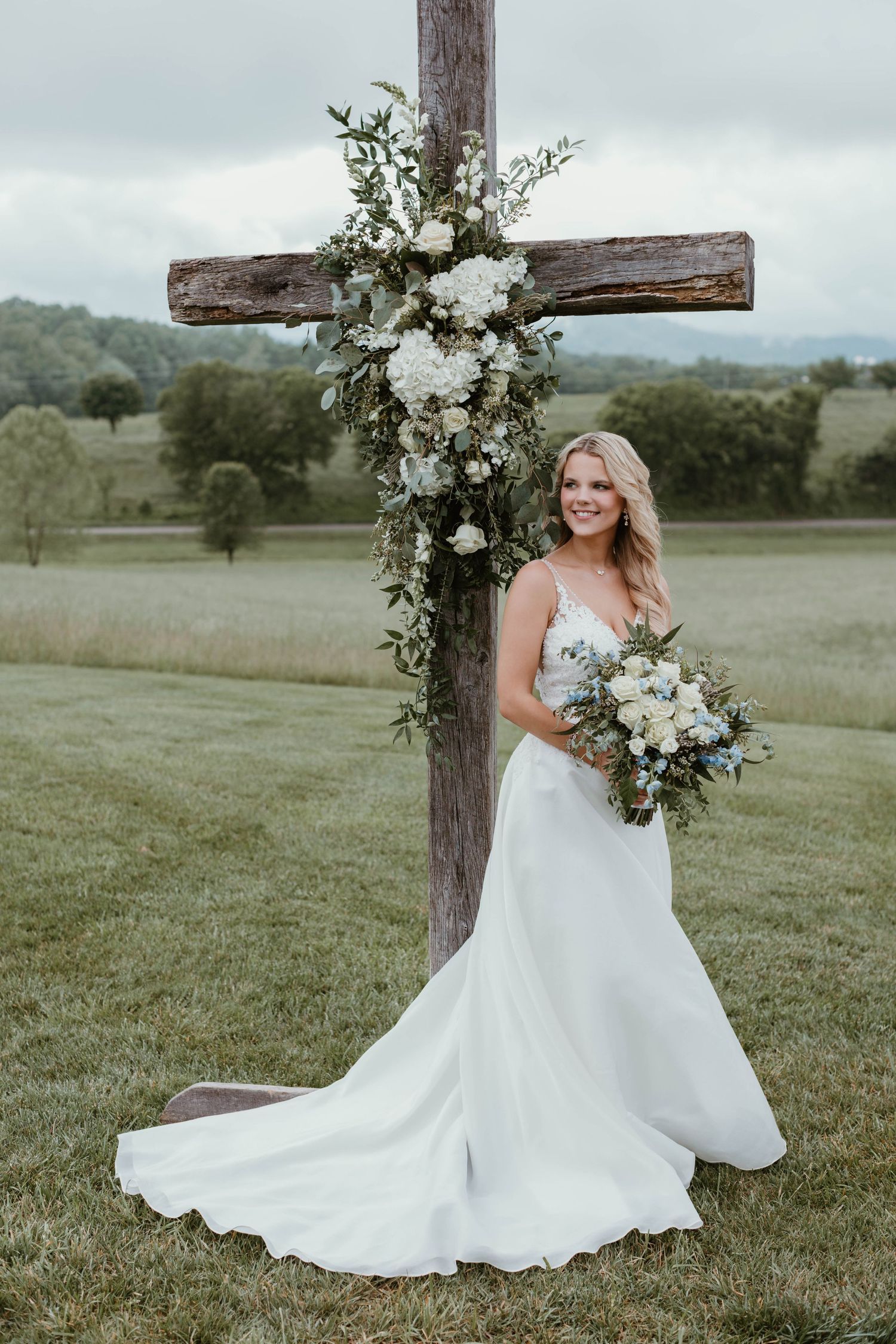 Flowing white wedding gown showcased in an outdoor field setting with rustic wooden cross.