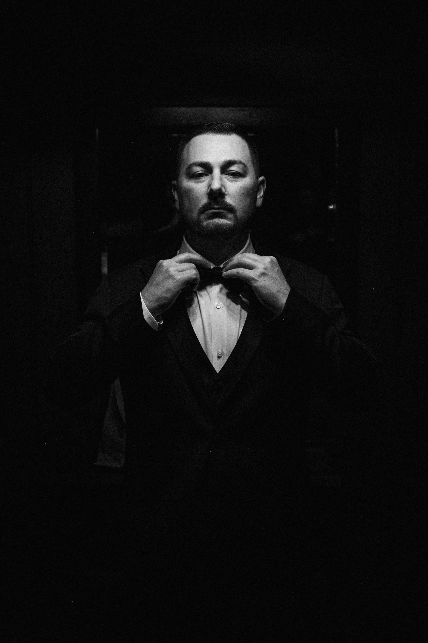 Black and white portrait of a man in a tuxedo adjusting his bow tie in dramatic low-key lighting.