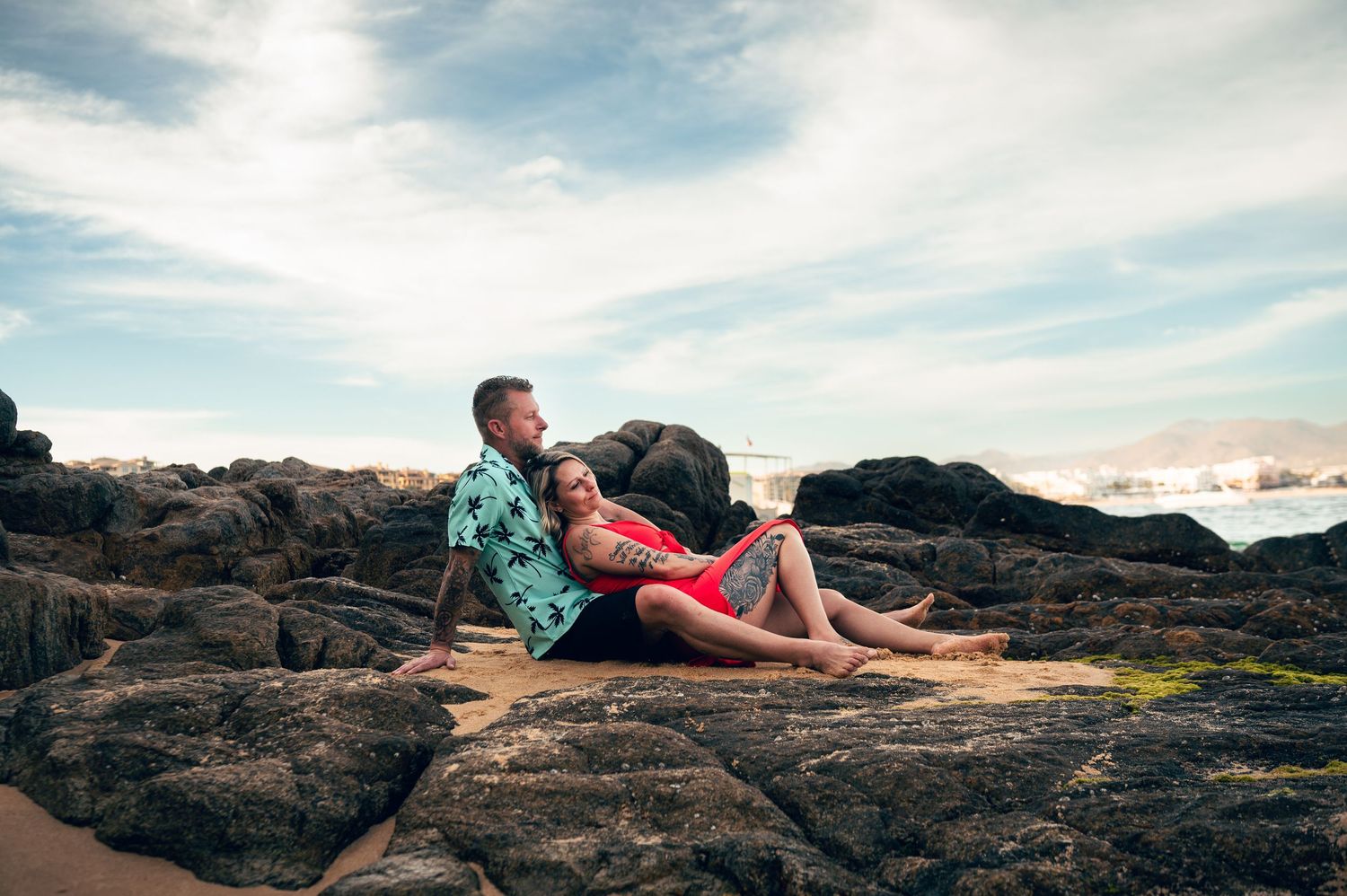 Couple sitting together on rocky beach shore, man in floral shirt and woman in red outfit, with ocean and cloudy sky behind.