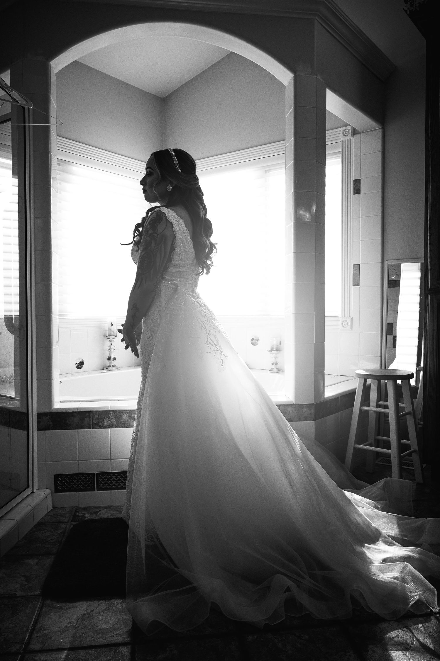 Bride in elegant white wedding gown with long train stands silhouetted by window light in black and white photo.