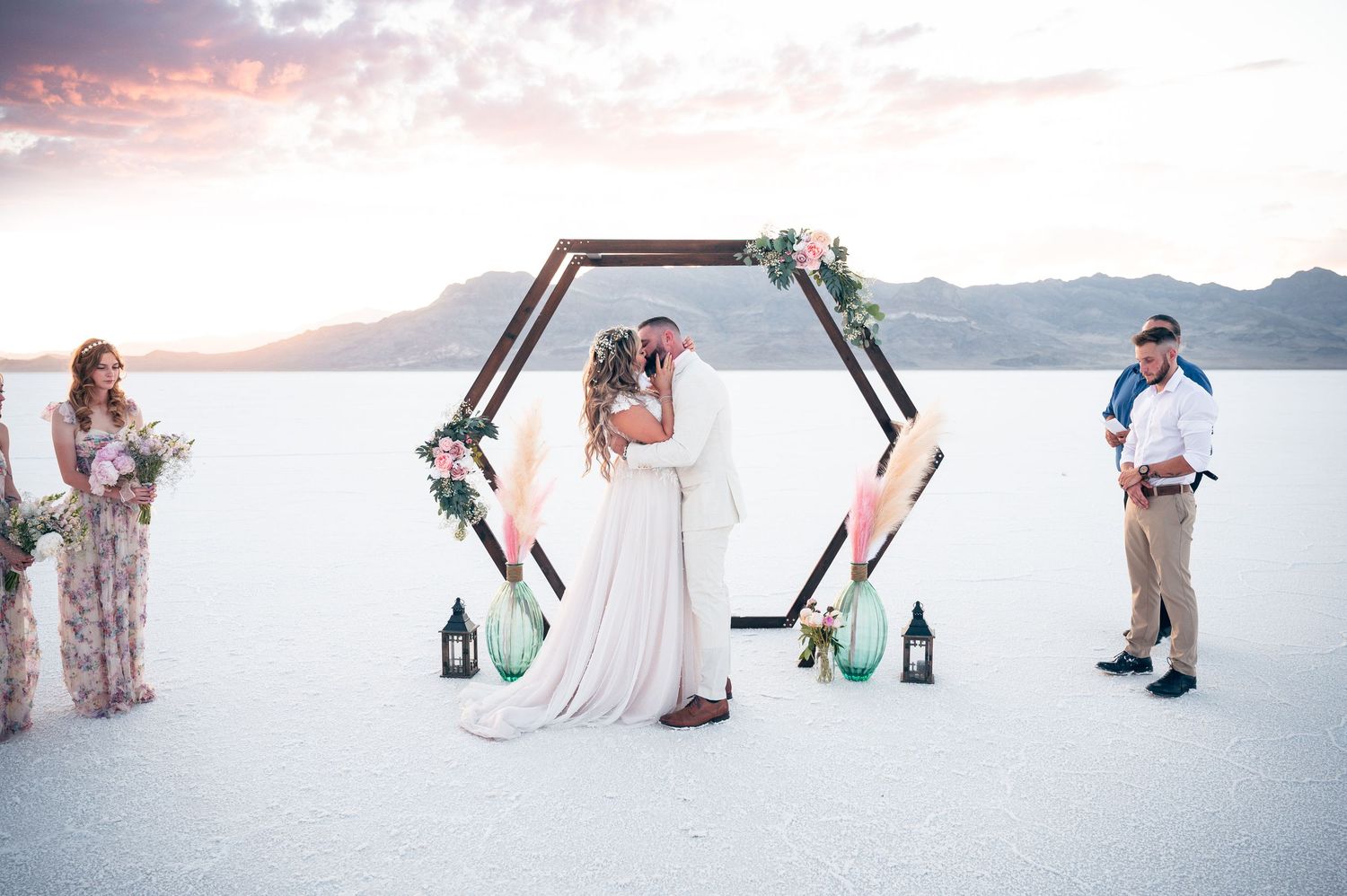 Couple kissing under hexagonal floral arch at outdoor wedding ceremony on white salt flats at sunset.