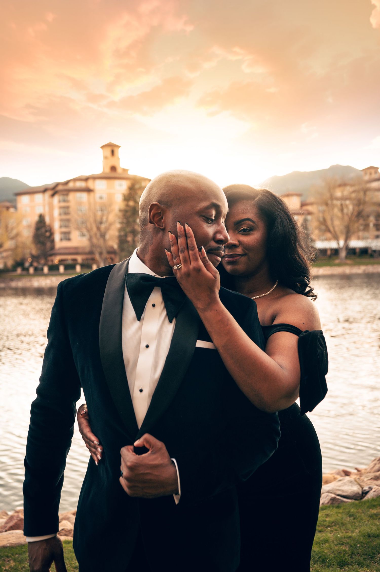 Elegant couple in formal attire embracing at sunset near a lake, woman in off-shoulder dress touching mans face tenderly.