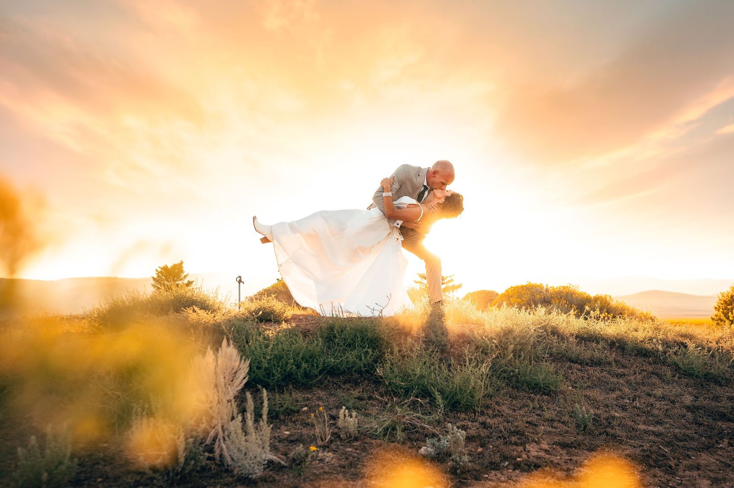 A couple shares a romantic dip kiss at sunset during their wedding on a scenic hilltop.
