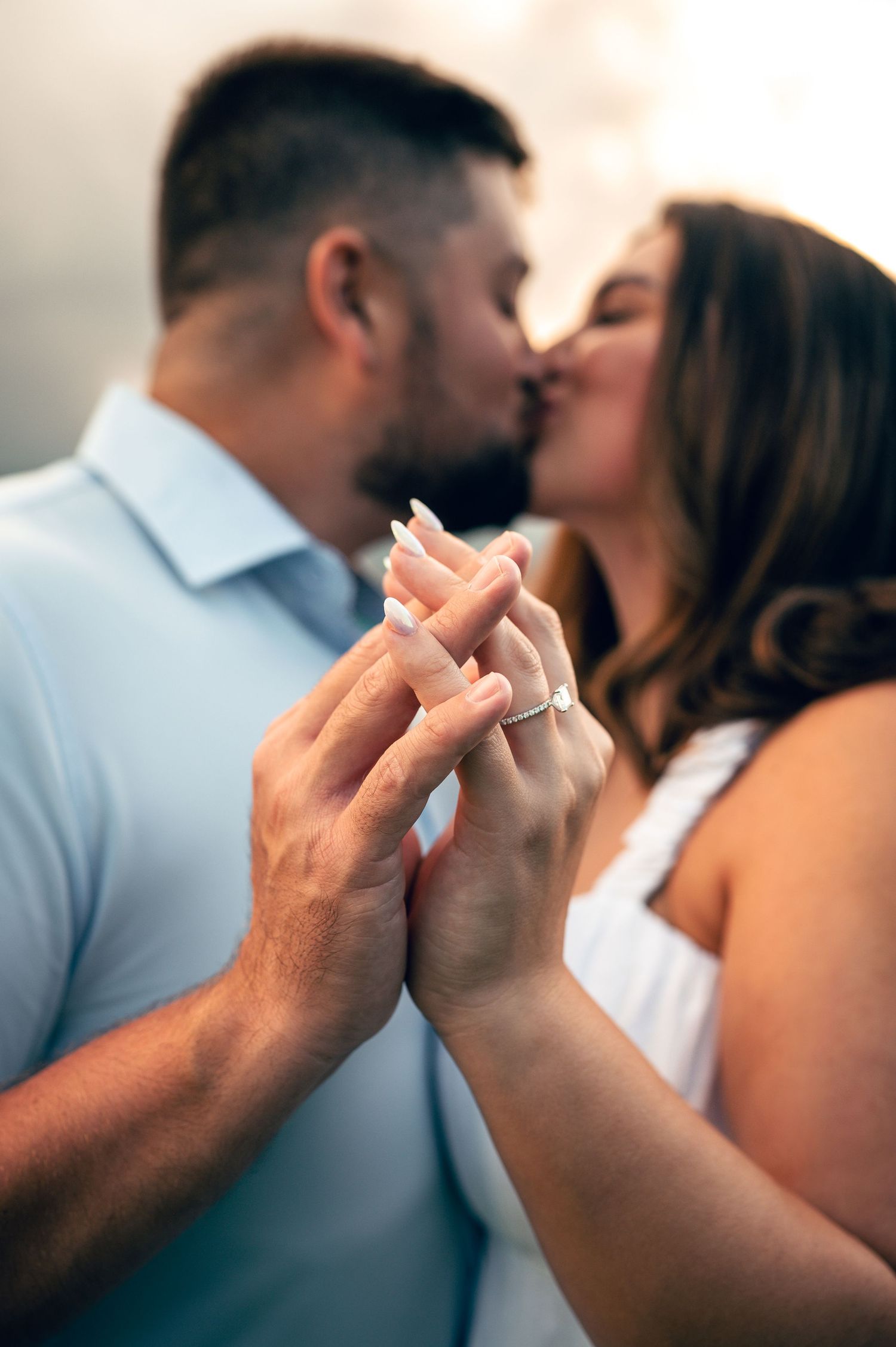 Couple sharing a romantic kiss while holding hands, showcasing an engagement ring, dressed in light blue and white.