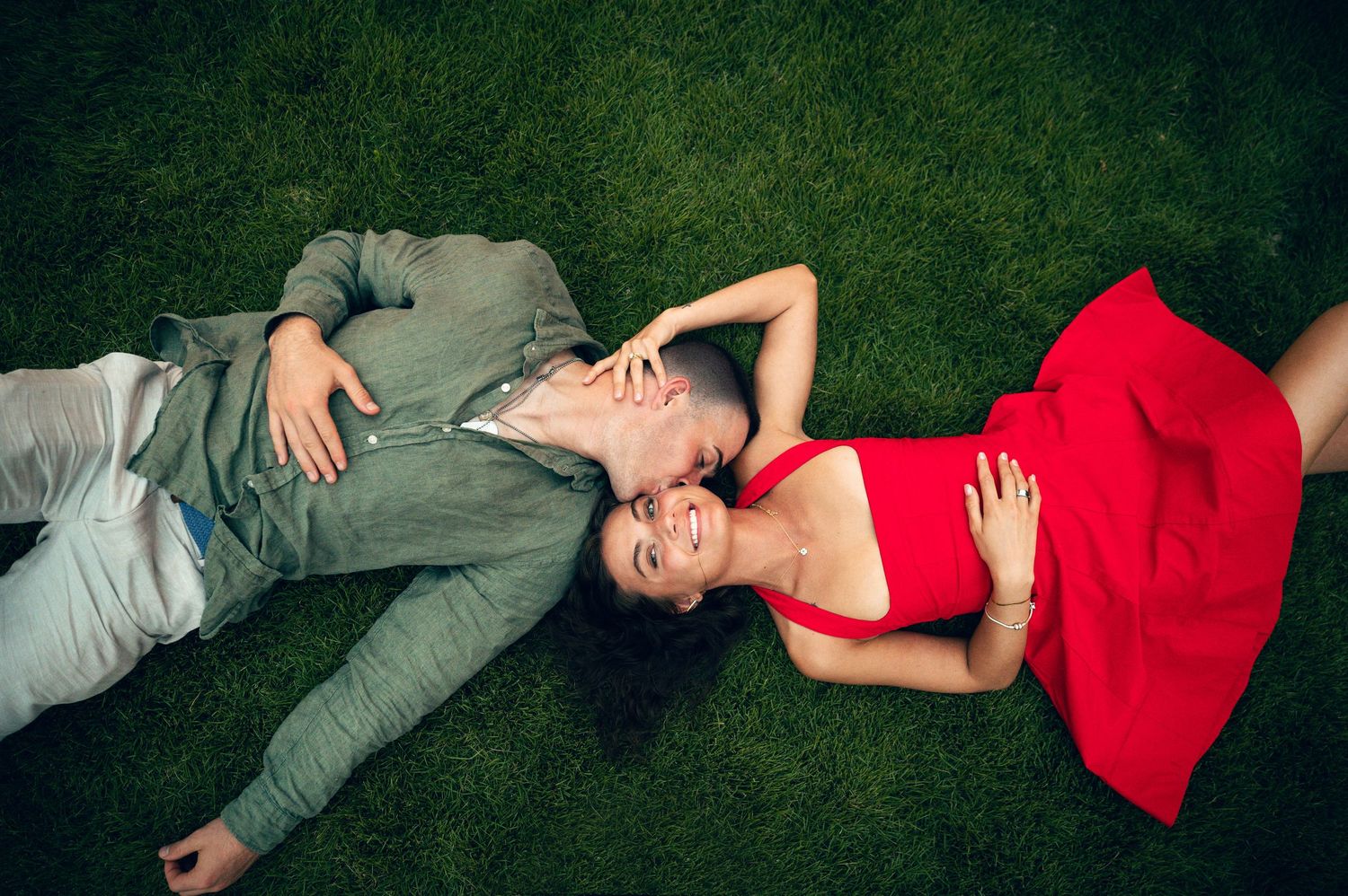Couple lying on green grass, man in gray suit kissing woman in red dress, romantic outdoor photo shoot.