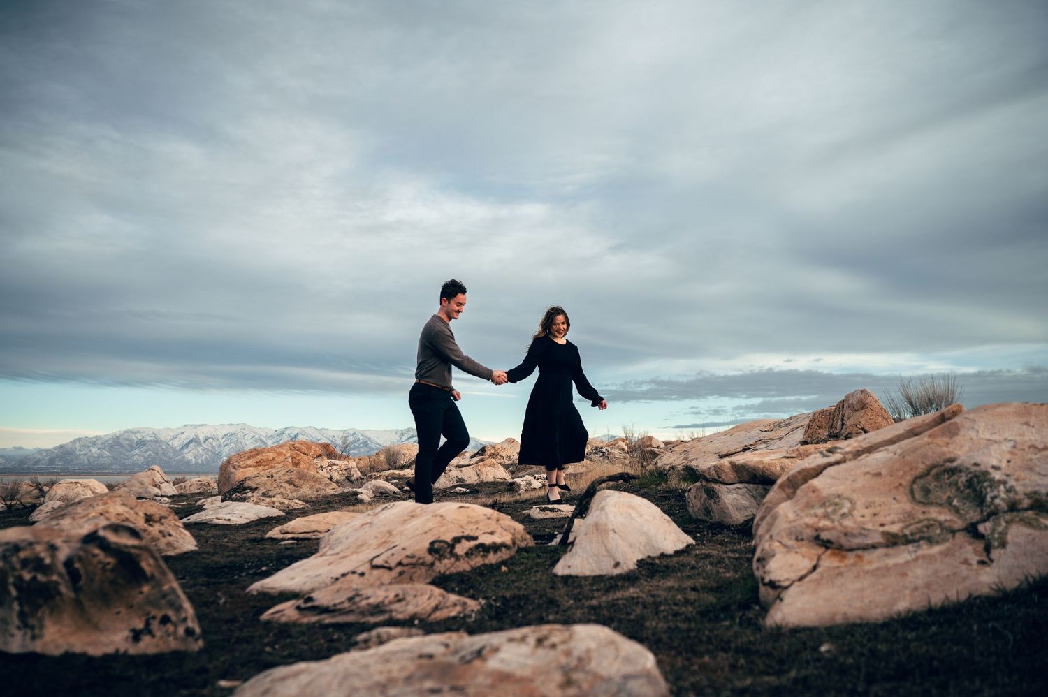 A couple holds hands while walking on rocky terrain with mountain views and dramatic cloudy skies behind them.