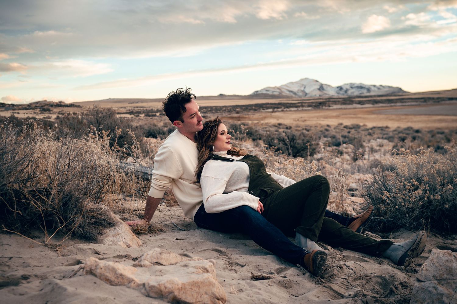 Couple sitting together on desert rocks at sunset with mountains and dramatic cloudy sky in background.