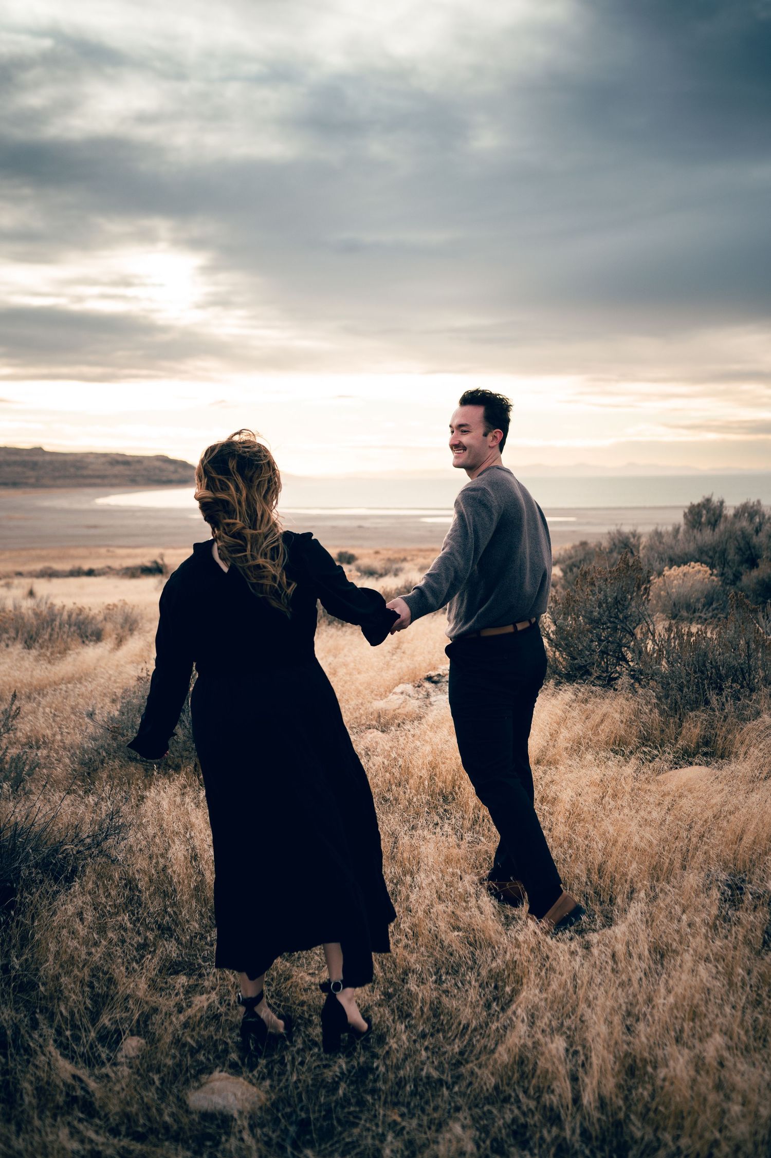 A couple holds hands and laughs while walking through golden grass fields near a coastal sunset.