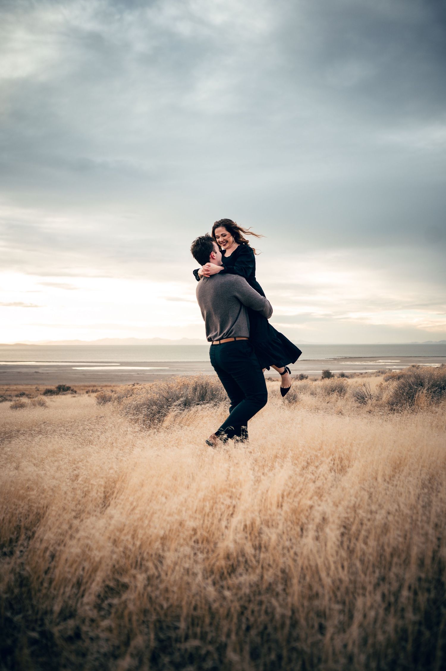 Couple embracing in golden coastal grasslands at sunset, man lifting woman as she laughs joyfully.